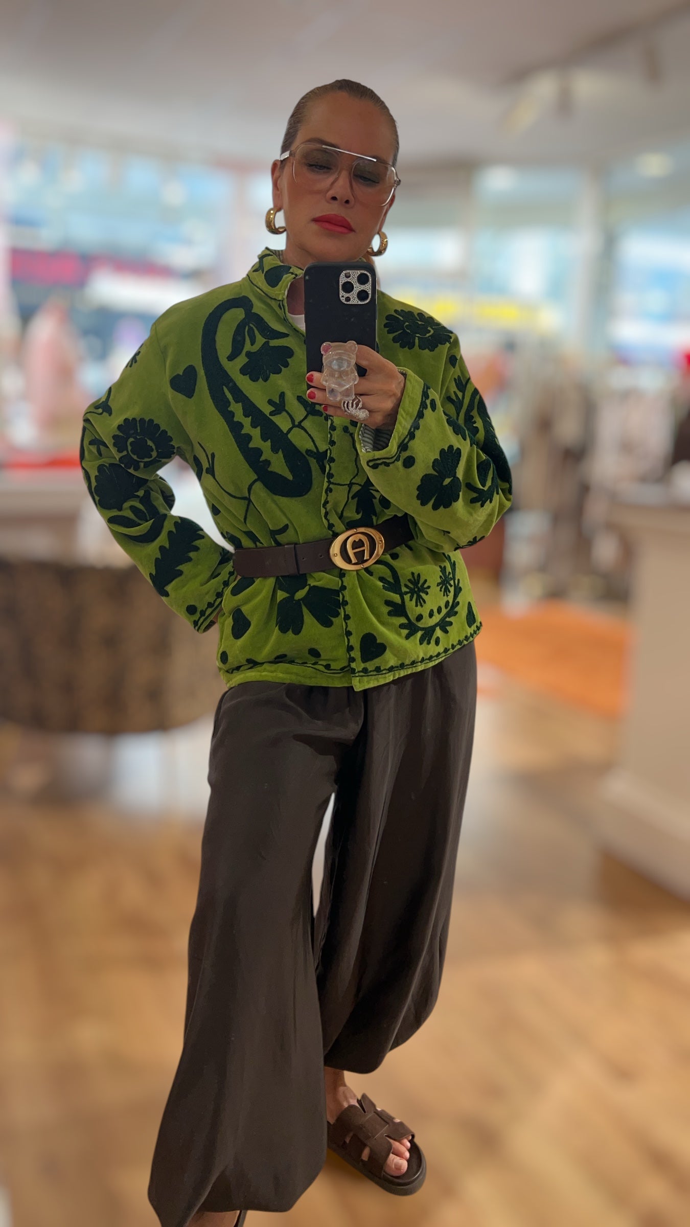 Frau trägt eine grüne gemusterte Jacke mit dunklem floralen Ornamentprint, Stehkragen und Knopfleiste. Kombiniert mit einer weiten schwarzen Hose und flachen Sandalen, fotografiert in einer hellen Boutique