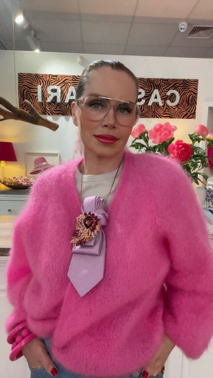 Frau trägt einen pinken Kid-Mohair-Cardigan mit heller Oberbekleidung, Jeans und dekorativem Accessoire im Brustbereich, stehend in einer Boutique.
