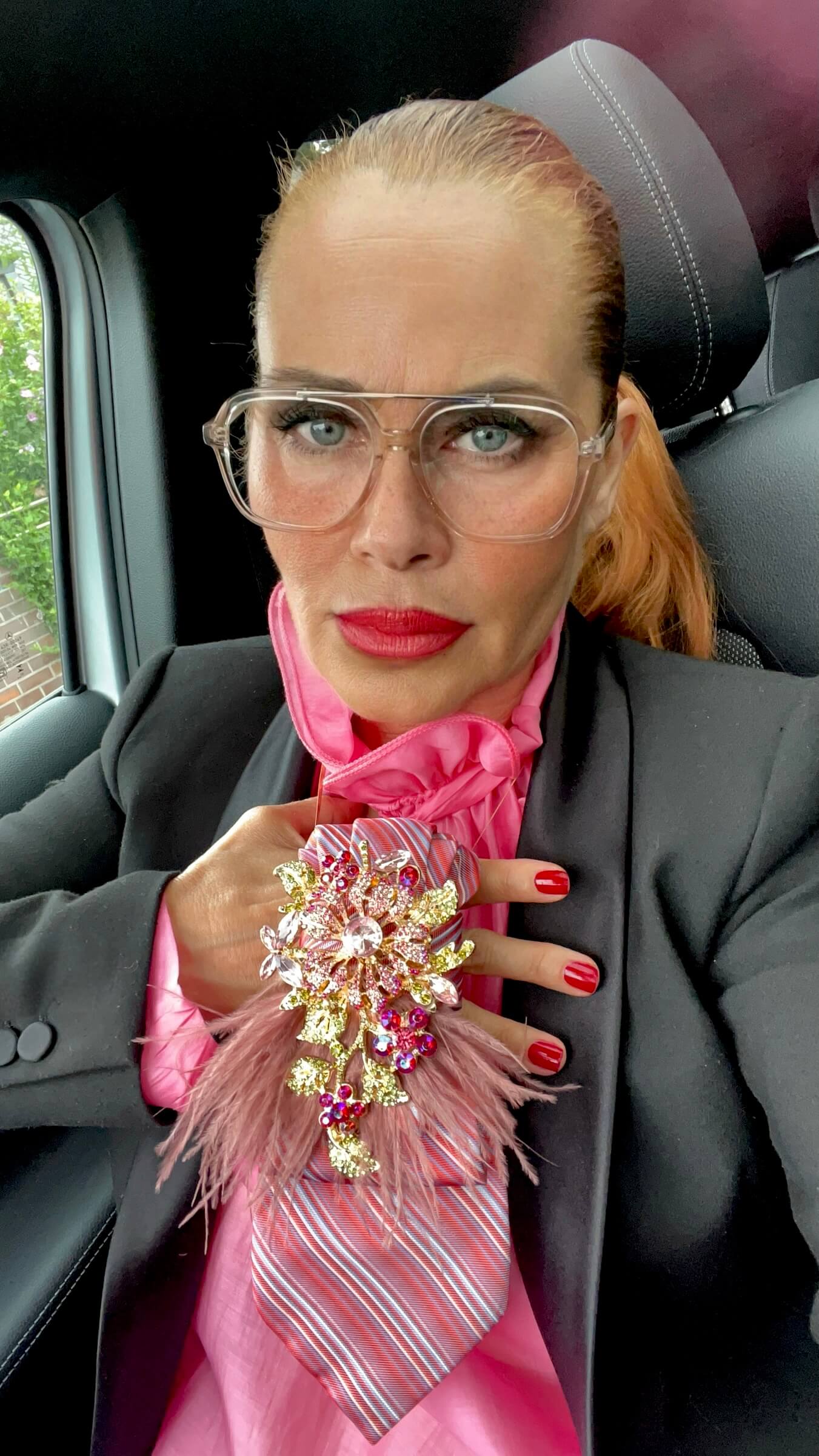 Frau mit schwarzem Blazer und pinker Bluse trägt eine rosafarbene Krawatte mit großer Brosche, Federdetails und Kette. Sie blickt direkt in die Kamera und hält die Krawatte mit einer Hand fest.