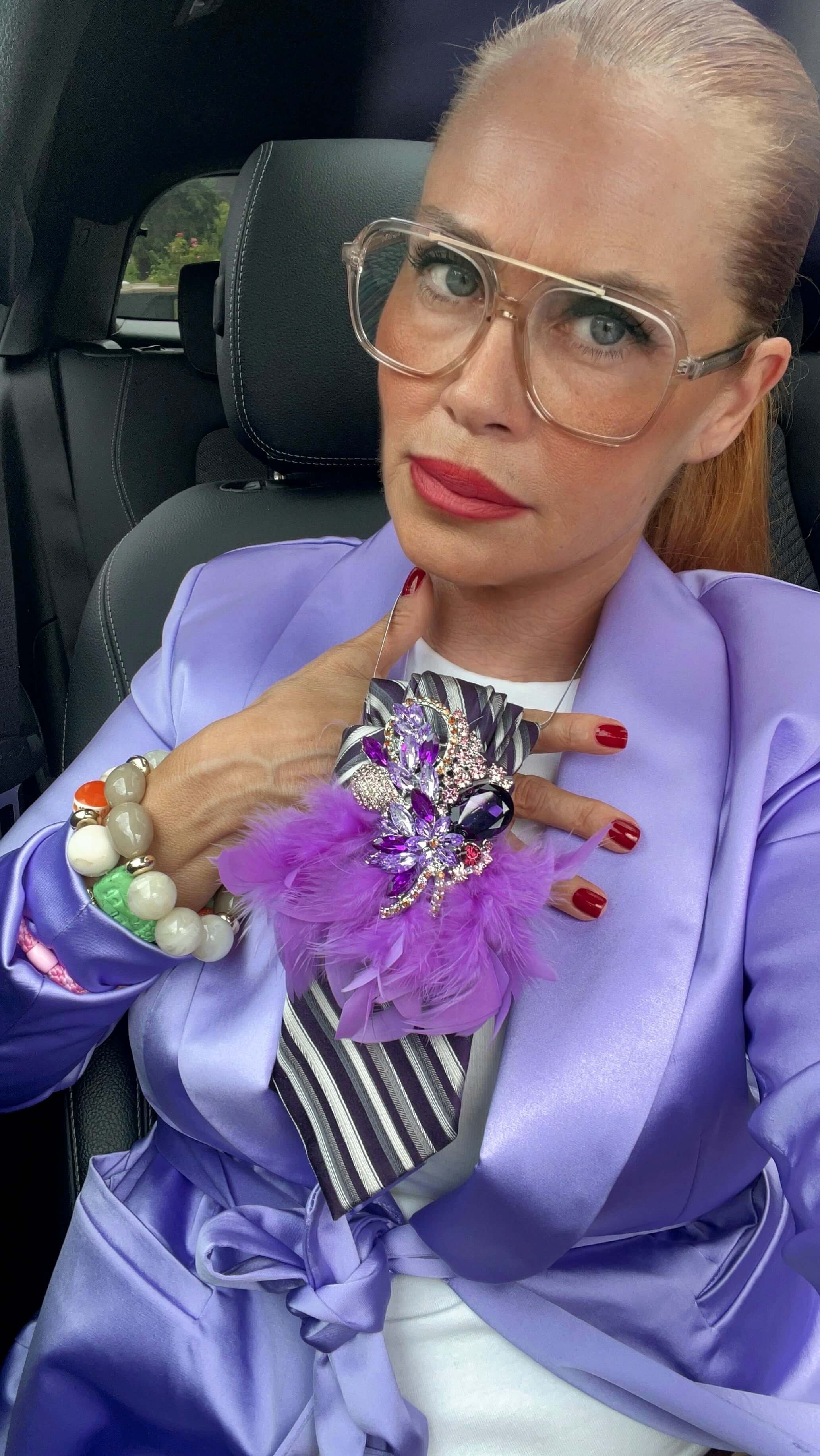 Frau trägt einen Lilabläser mit einer lila Krawatte Federn und einer großen Bronchitis sitzt im Auto und macht ein Selfie