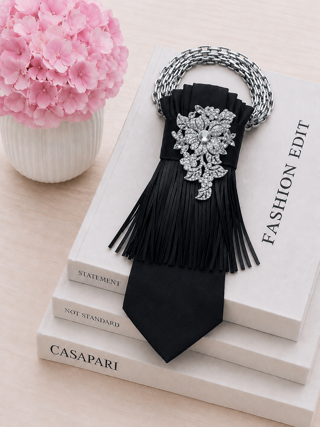 Schwarze Statement-Krawatte mit silberfarbener Gliederkette, funkelnder silberner Blütenbrosche und feinen Fransen auf dekorativen Fashion-Büchern mit rosa Hortensien.