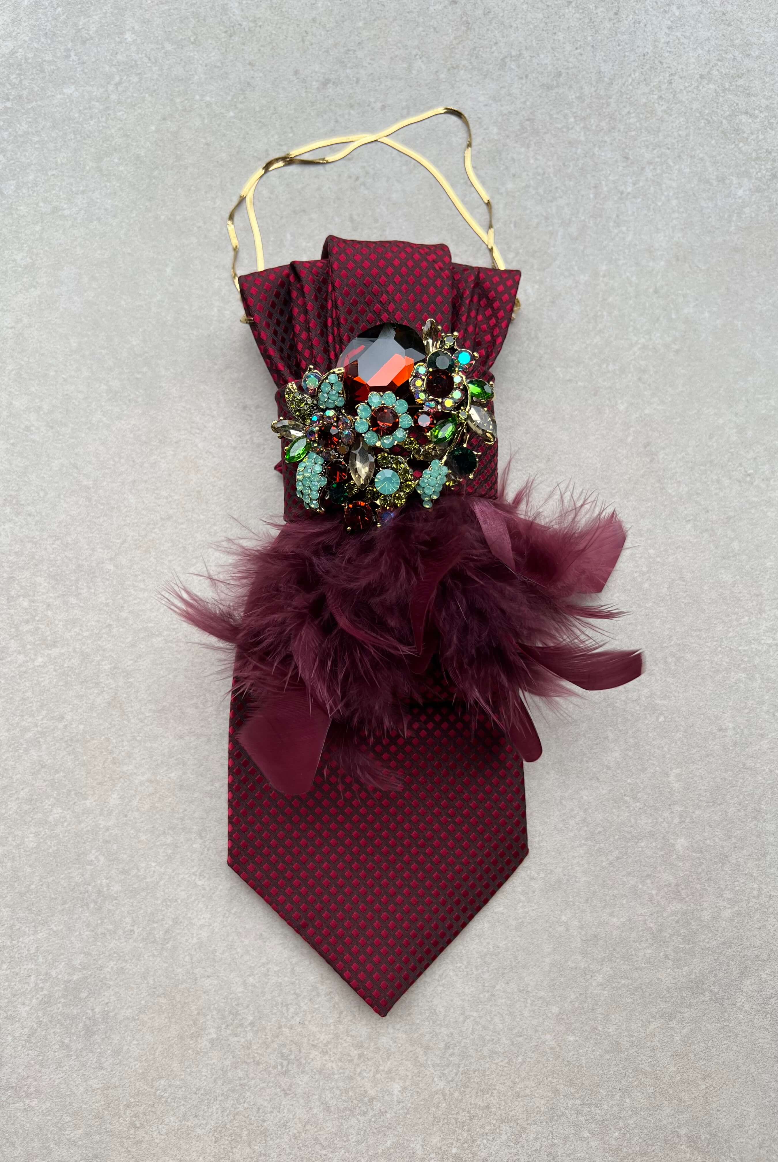 Bordeauxrote Feather-Affair-Krawatte mit feinem Muster, goldfarbener Kette, opulenter Kristallbrosche in Rot, Türkis und Grün sowie bordeauxfarbenen Federn