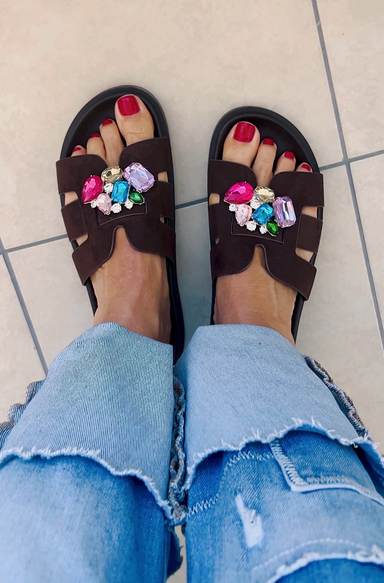 Braune Sandalen mit aufgesteckten Schuhclippern aus mehrfarbigen Schmucksteinen, getragen zu hellblauer Jeans.