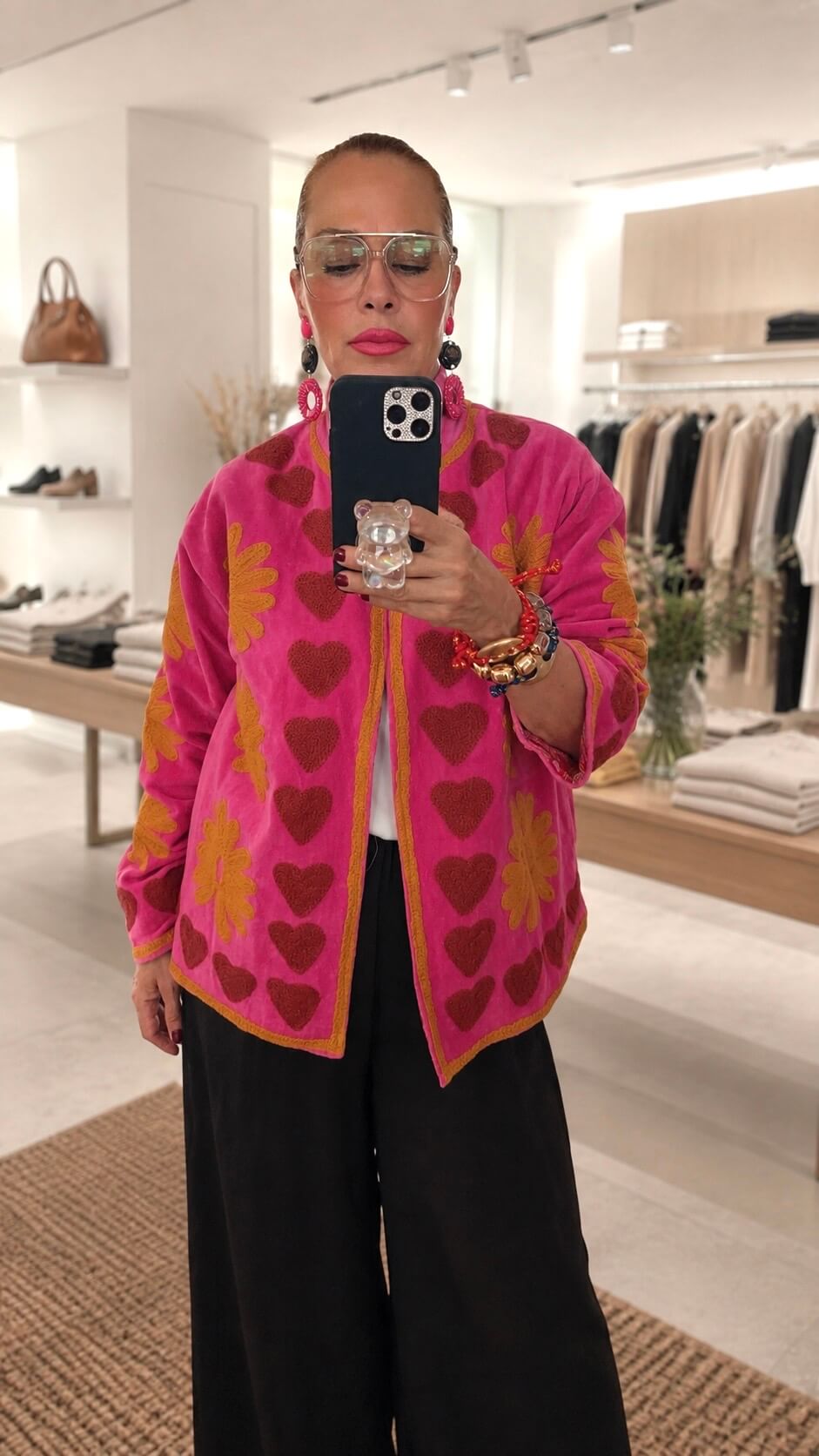 Frau steht in einer Boutique. Sie hat eine pinke Jacke mit Herzen an und macht ein Selfie
