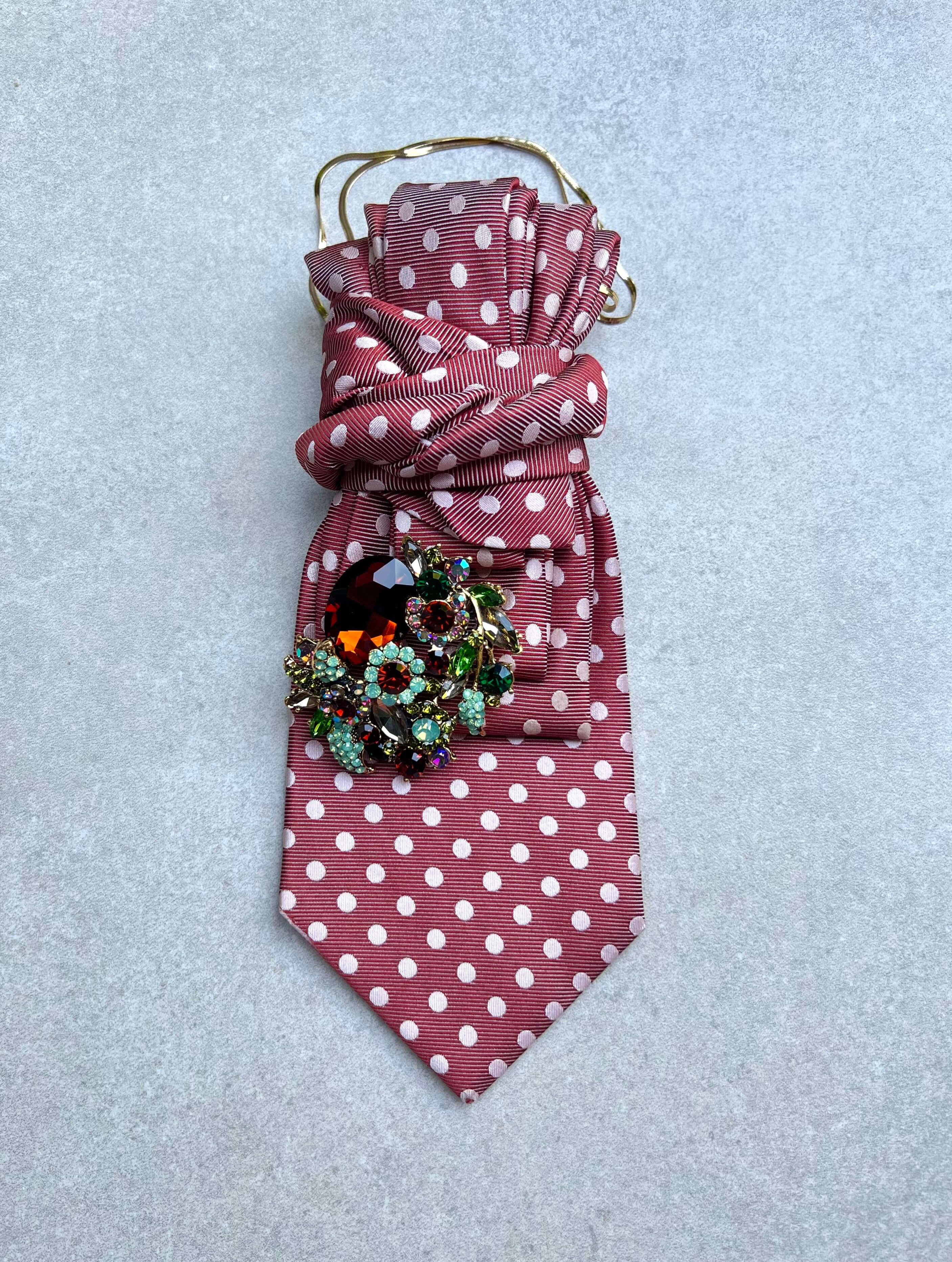 Rosafarbene Vintage-Krawatte mit weißen Punkten, goldfarbener Kette und großer Schmuckbrosche mit roten, grünen und türkisfarbenen Kristallsteinen. Modell Rose Society von Casapari.