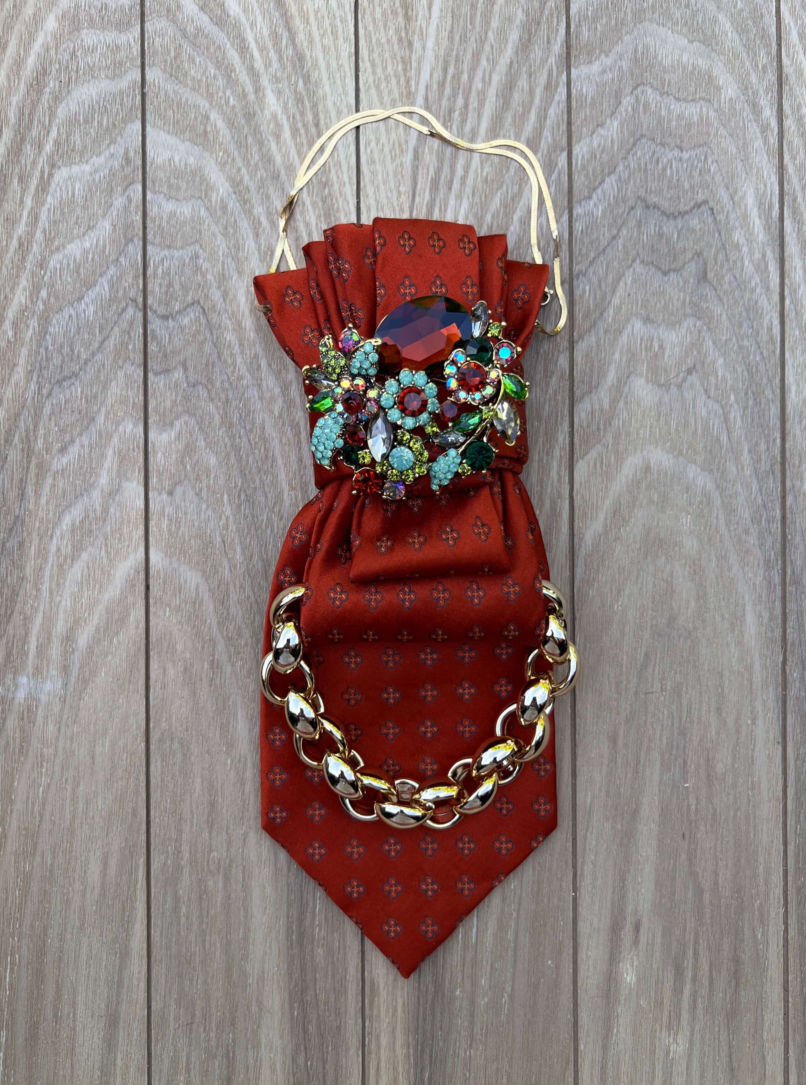 Rote Vintage-Krawatte mit Kristallbrosche und goldfarbener Kette auf hellem Holzboden arrangiert.