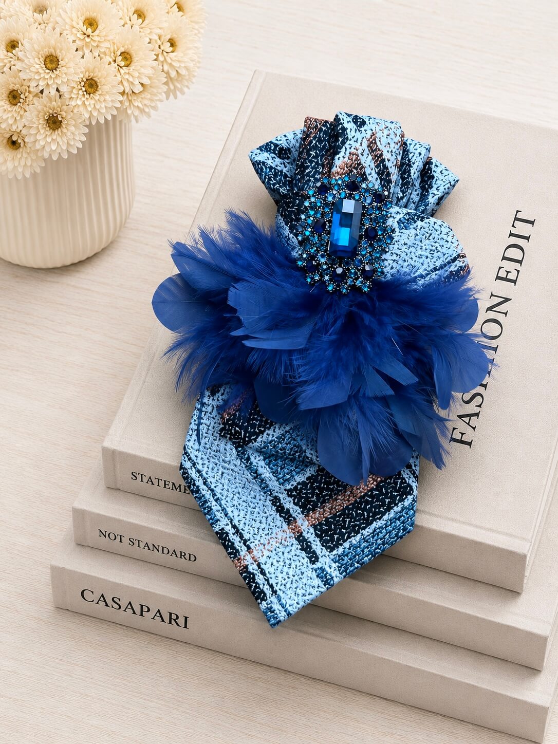 Blaue Vintage-Seidenkrawatte mit Schmuckbrosche und dunkelblauen Federn auf cremefarbenen Designbüchern arrangiert.
