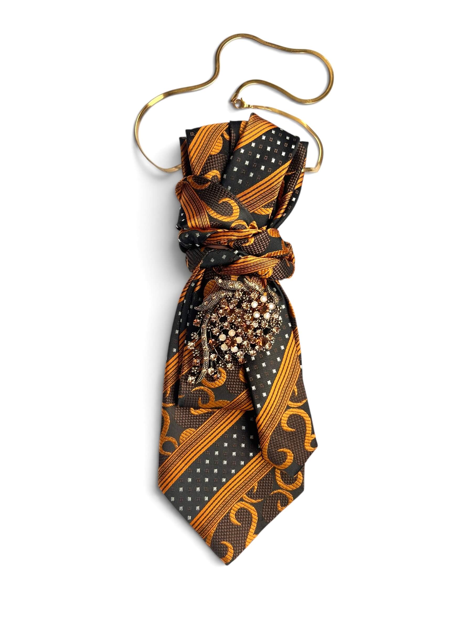 Krawatte in warmem Amber und Schwarz mit Muster, mittig gebunden und mit Brosche verziert, vor hellem Hintergrund.