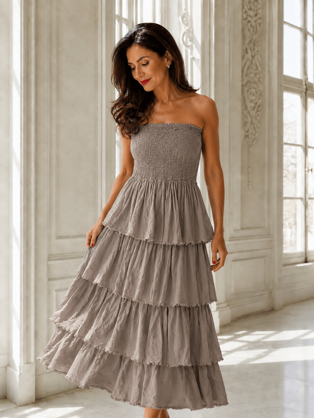 Frau trägt ein taupefarbenes Boho-Kleid aus reiner Viskose mit elastischem Smok-Oberteil und mehrlagigem Volantrock in einem hellen, eleganten Interieur.