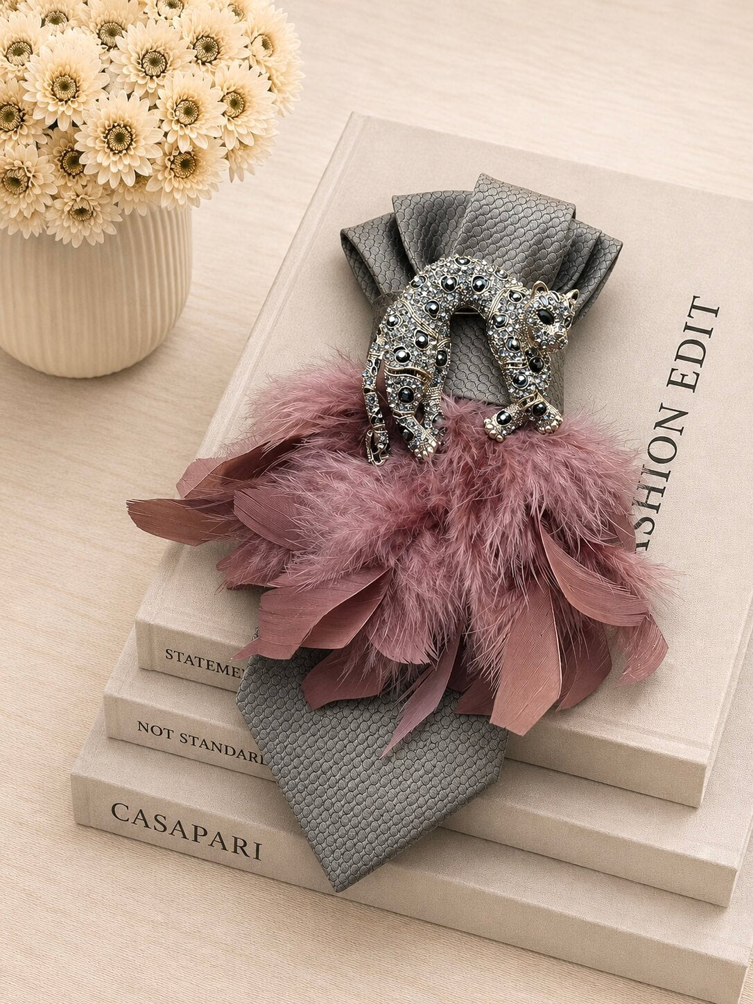 Graue Vintage-Seidenkrawatte mit Leopardenbrosche und roséfarbenen Federn auf cremefarbenen Designbüchern arrangiert.