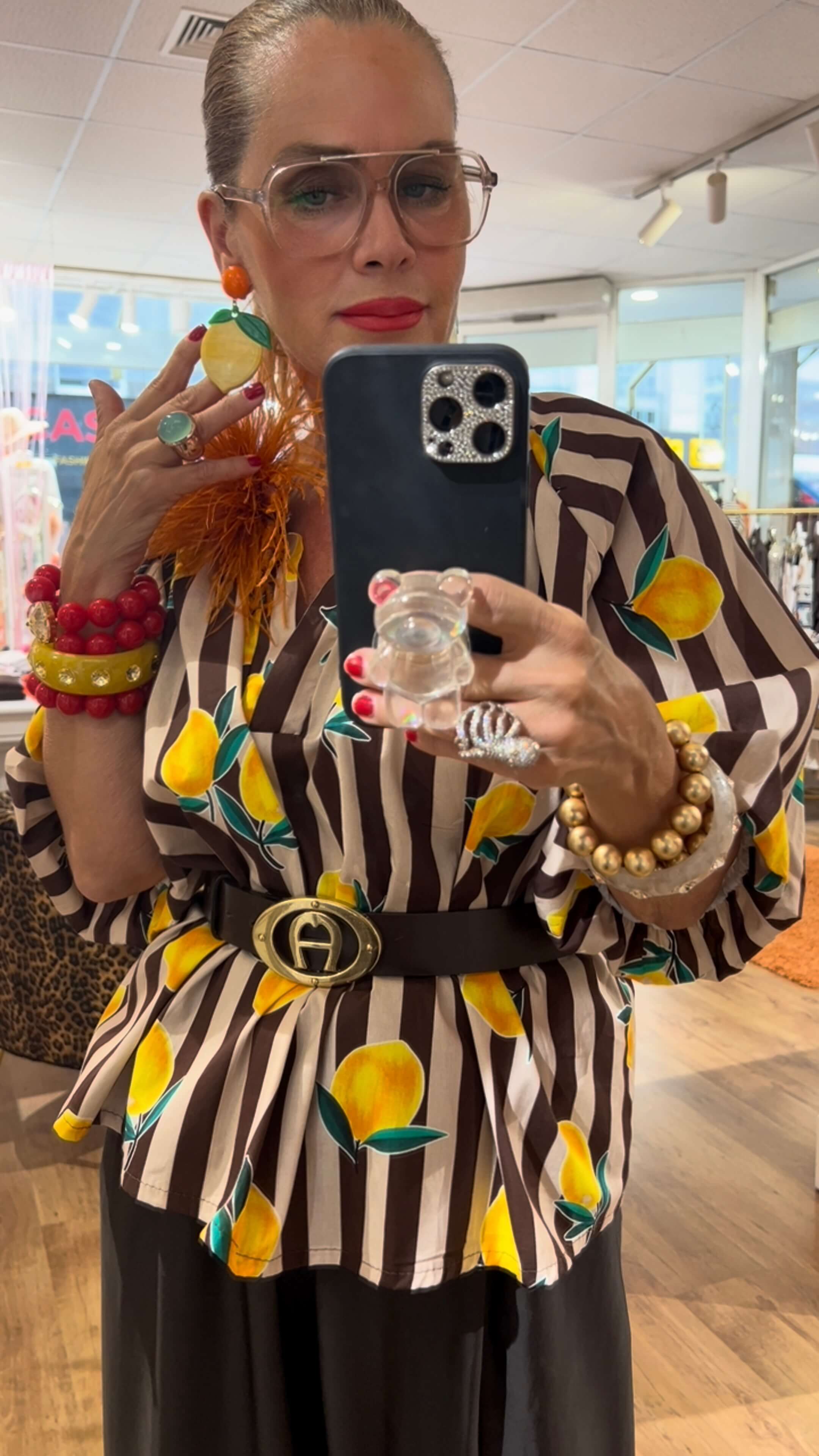 Frau mit Zitronen-Ohrringen, gestreifter Zitronen-Bluse, Gürtel und auffälligem Armschmuck vor einem Spiegel im Store.