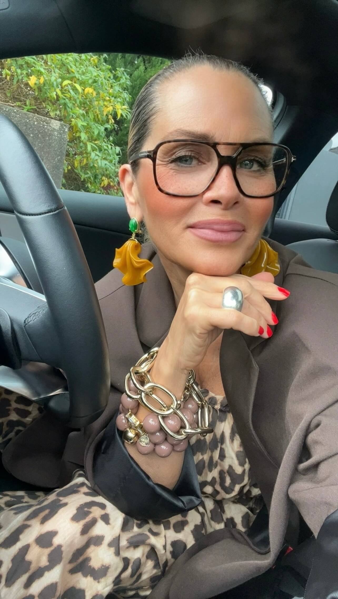 Frau mit großer dunkler Brille, sitzt im Auto. Sie trägt einen großen silbernen Ring, große Ohrringe. Ein Leo Kleid und braunen Blazer.