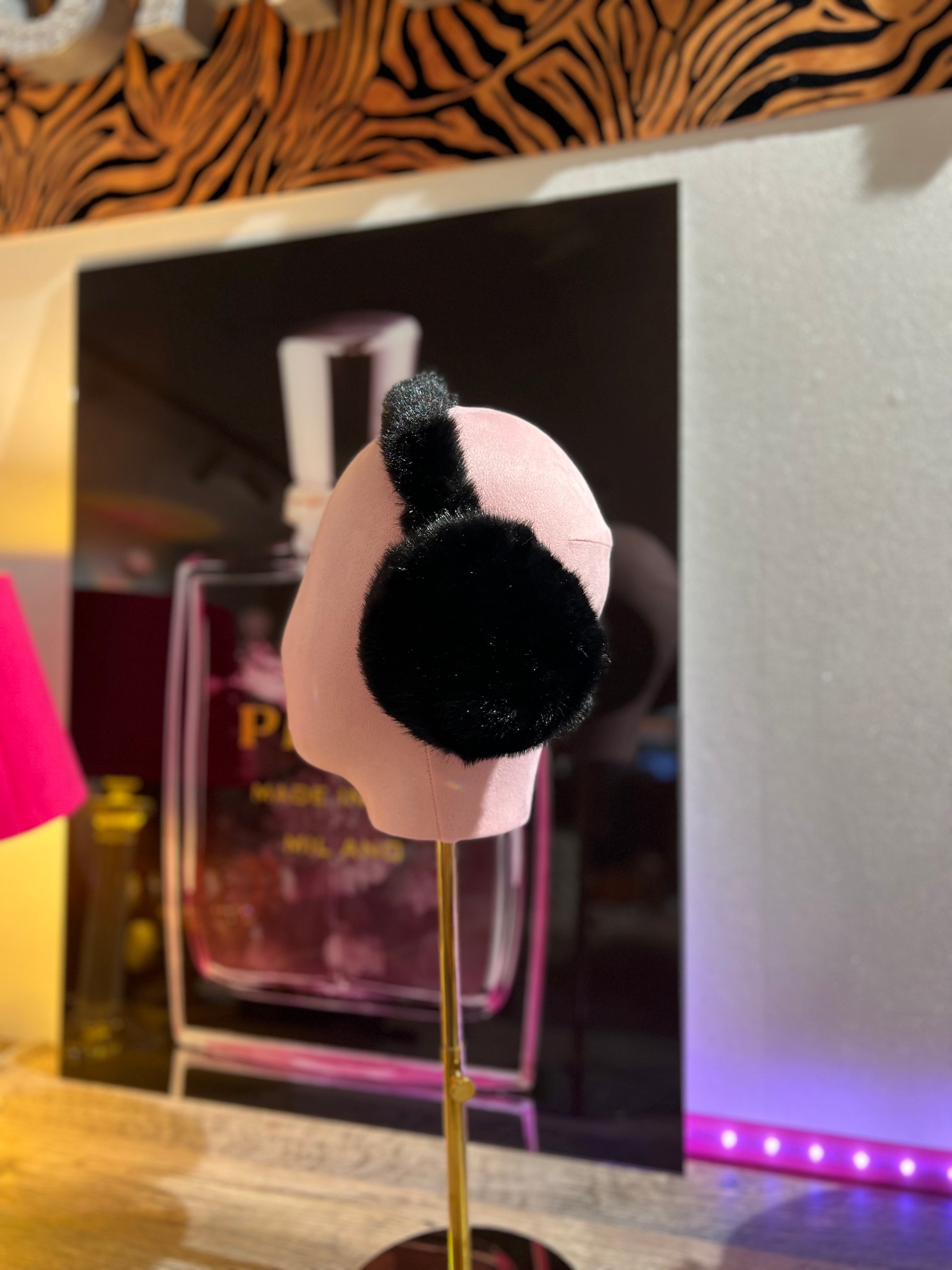 Schwarze, flauschige Ohrwärmer auf rosafarbene Präsentation Büste, im Hintergrund unscharf ein Parfumflakon und warme Beleuchtung.