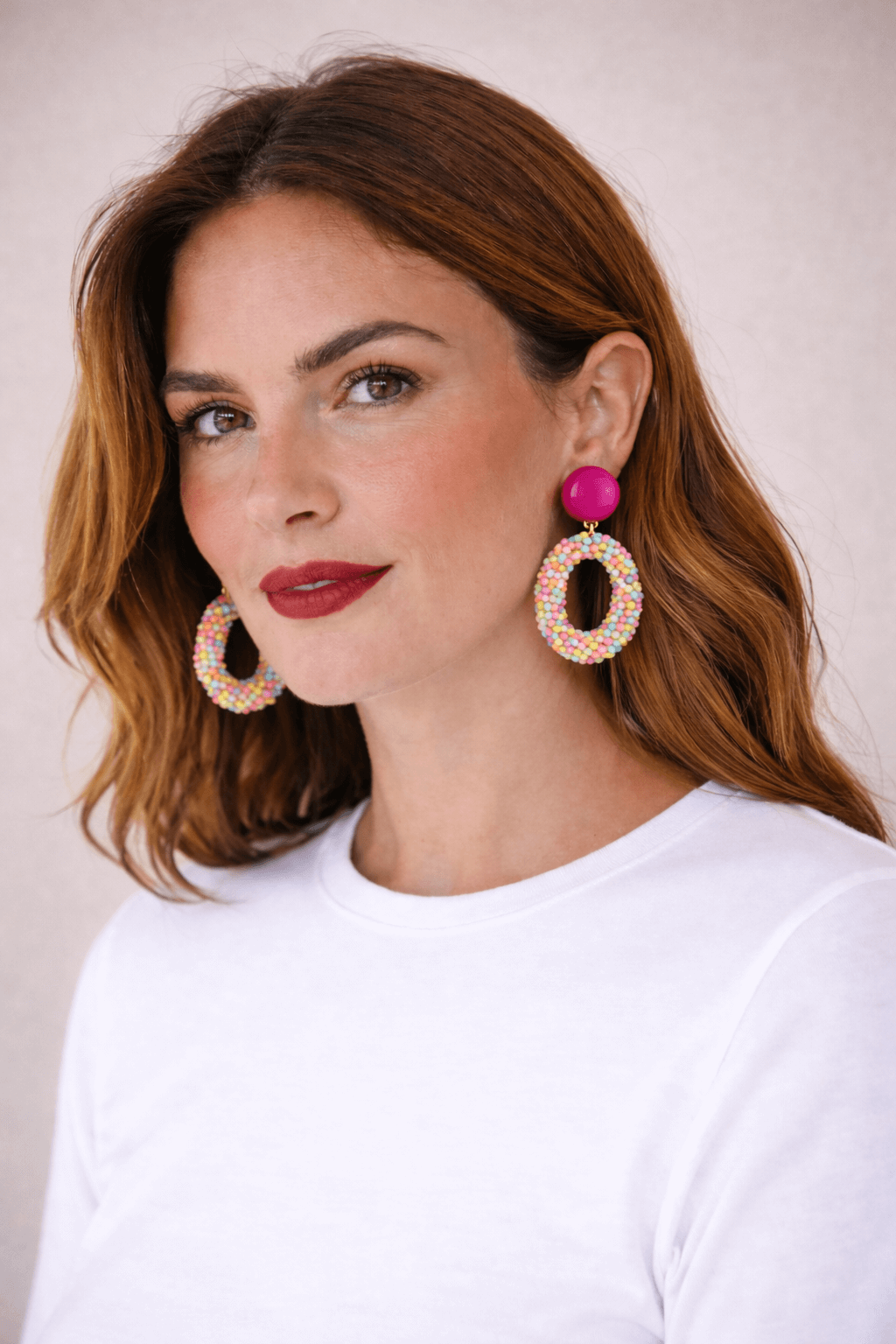 Frau mit schulterlangem braunem Haar trägt ovale Ohrringe mit pinkem Clip-Verschluss und bunten Perlen in Pastelltönen vor hellem Hintergrund.