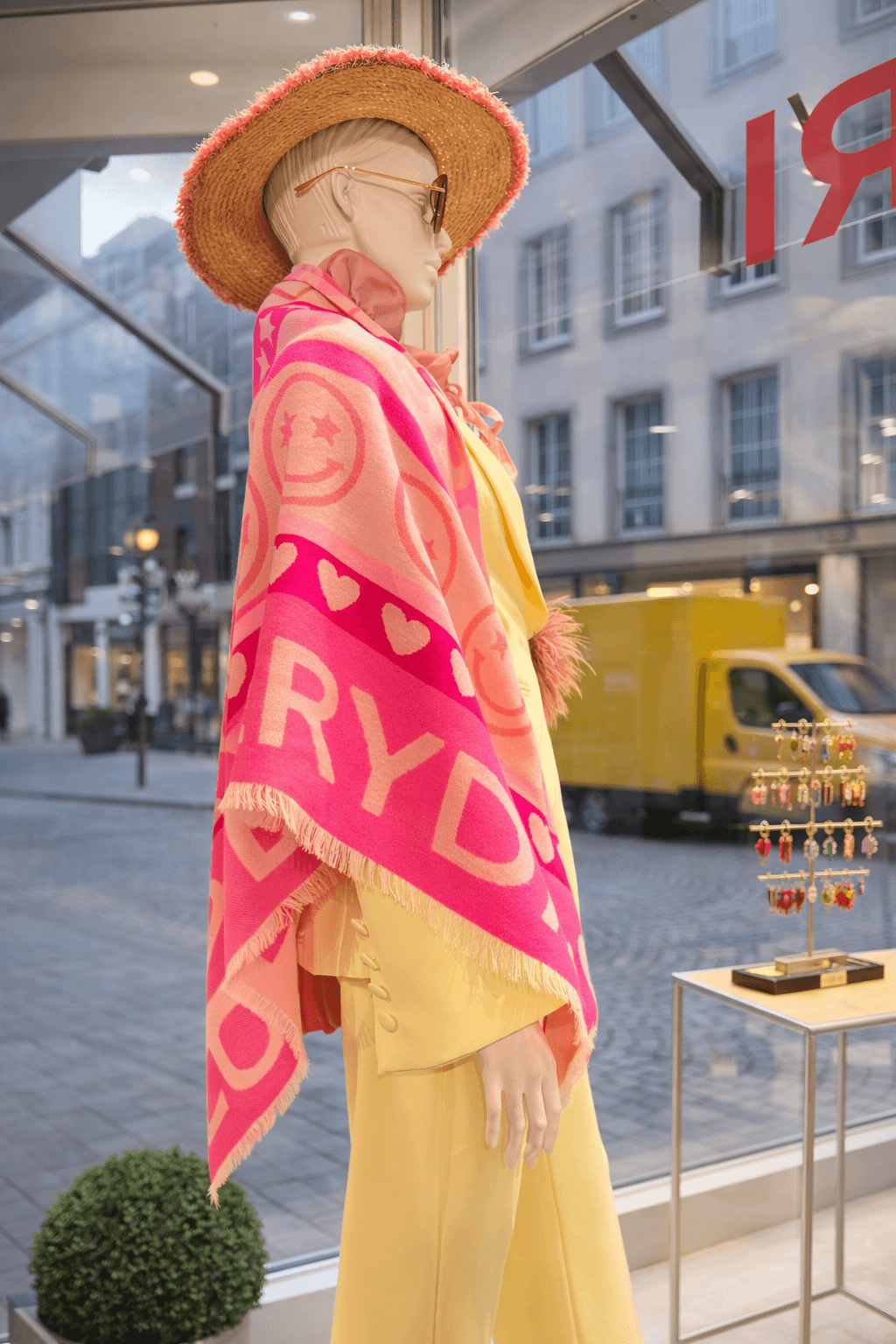 Schaufensterpuppe im Profil trägt pink-rosafarbenen Poncho mit Schriftzug, gelbe Hose, Strohhut und Sonnenbrille. Hinter der Schaufensterscheibe ist eine Straße mit geparkten Fahrzeugen und Gebäudefassade zu sehen.
