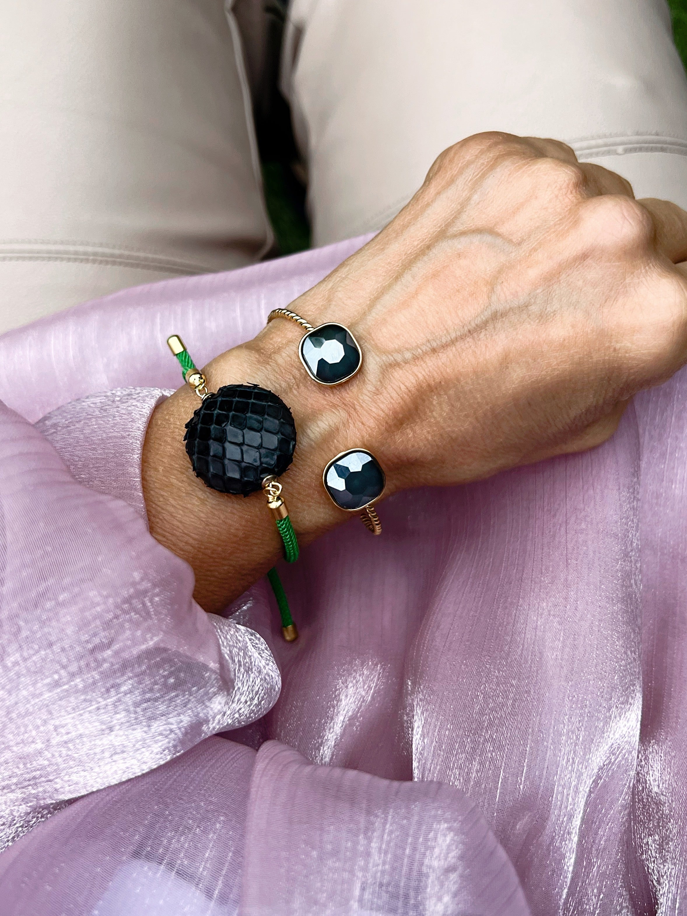 Ein grünes Armband mit schwarzer Perle dazu ein goldener Armreif mit  Glasstein in Taupe. An einem Handgelenk auf rosa Stoff fotografiert.