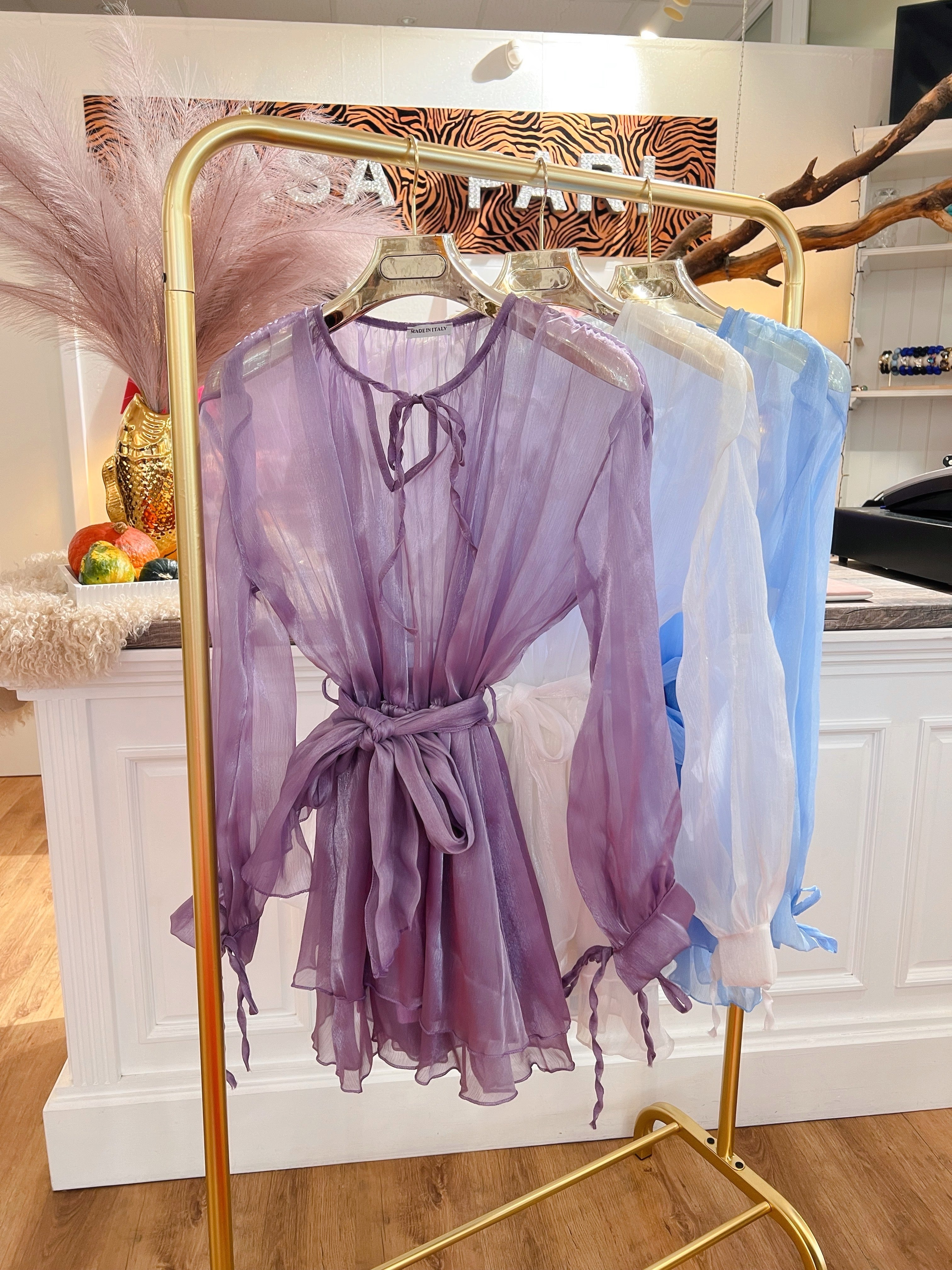 Mehrere transparente Blusen in Flieder, Weiß und Hellblau hängen auf goldenen Kleiderbügeln an einem goldenen Kleiderständer vor weißen Theke im Laden.