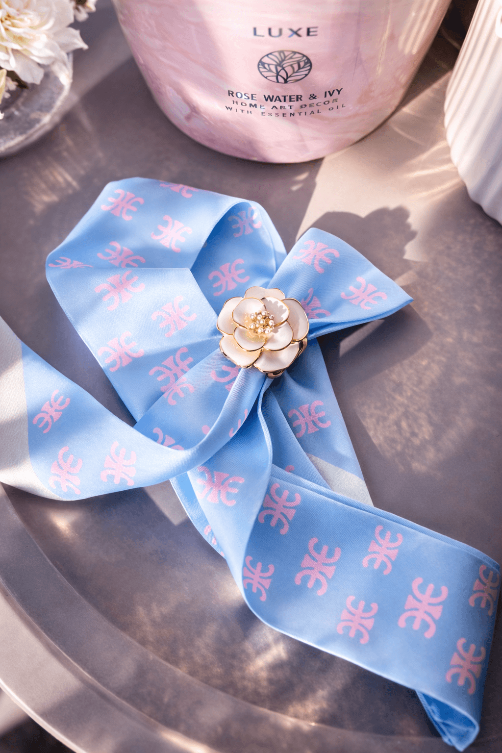 Hellblauer Schal mit rosafarbenem Muster liegt auf runder Tischplatte, fixiert mit goldfarbenem Schalring in weißer Blütenform mit Perlendetail; im Hintergrund rosa Kerze und weiße Vase.