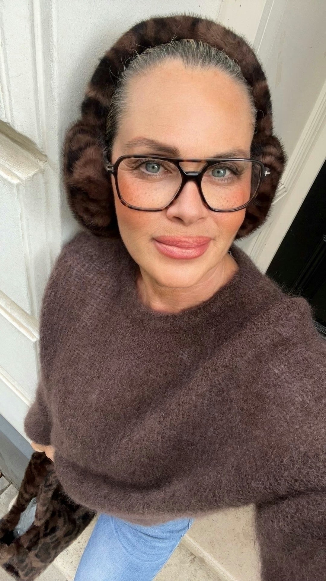 Frau trägt braune Ohren Ohrenwärmer in Leo. Dazu brauner Mohairpulli. Fotografiert an einem hellen Haus.