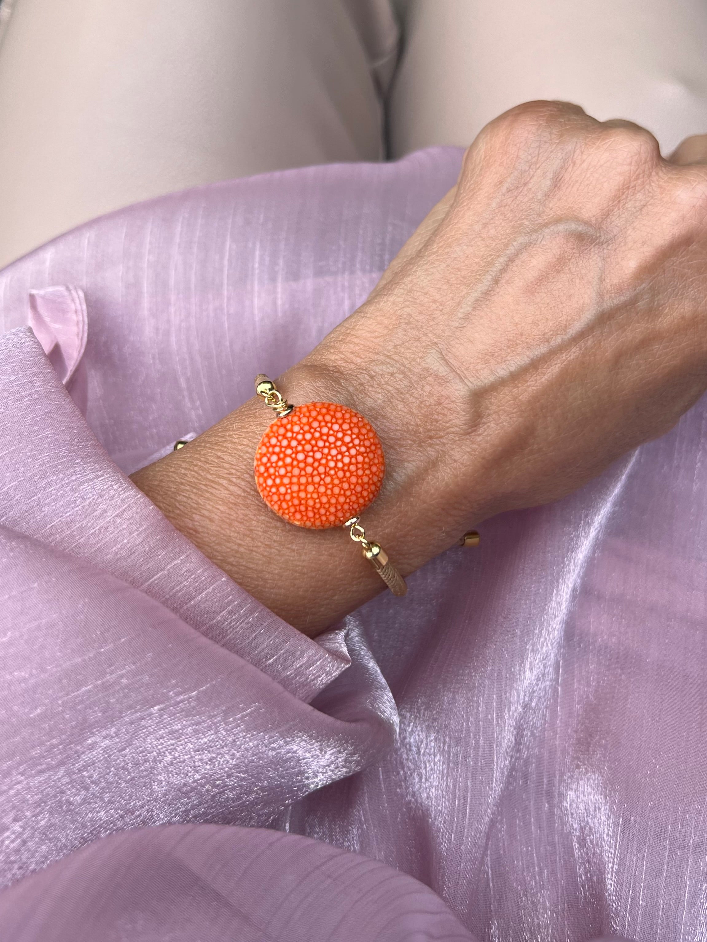 Beiges Armband mit oranger Perle und goldene Details. Am Handgelenk auf rosa Stoff fotografiert.