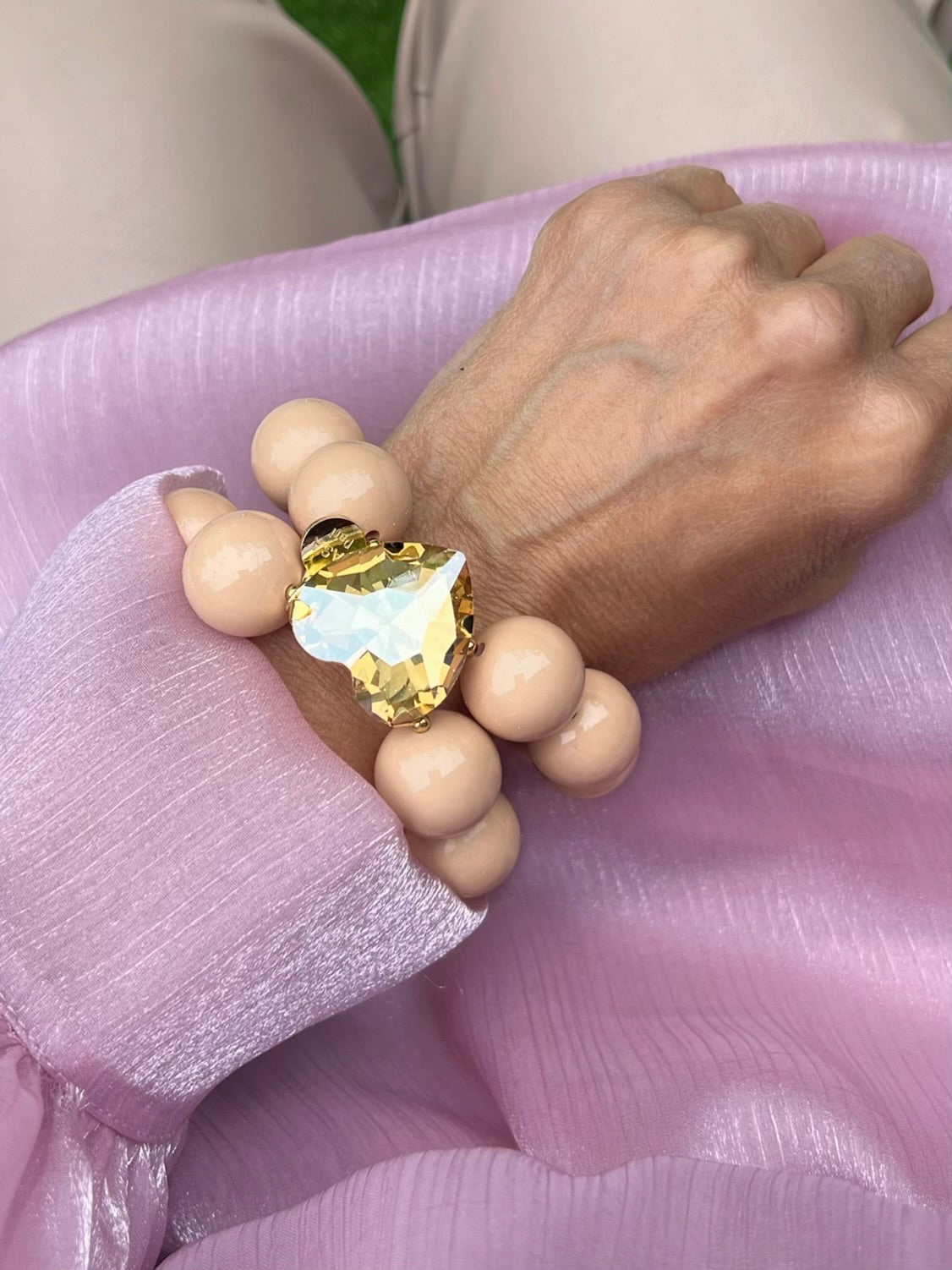Armband in Hellbeige mit großem Herzschmuckstein. Am Handgelenk auf rosa Stoff fotografiert.