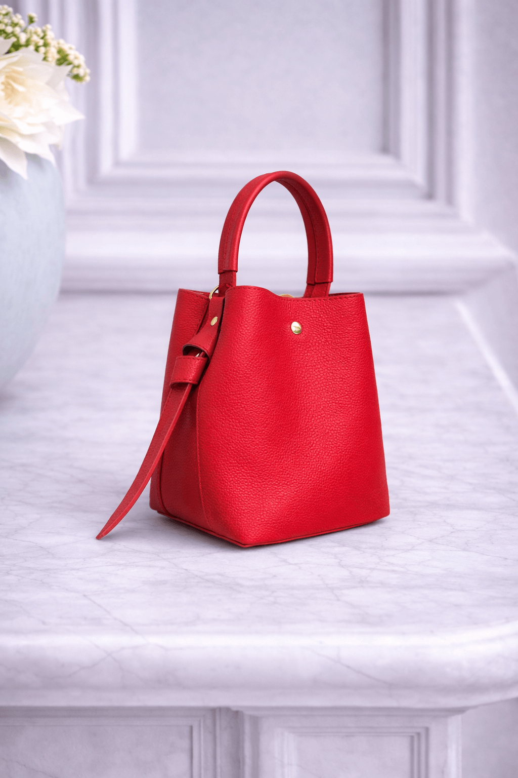 Kompakte Handtasche aus rotem Nappaleder mit rundem Henkel und goldfarbener Niete, stehend auf einer hellen Marmorfläche vor ruhigem Hintergrund.
