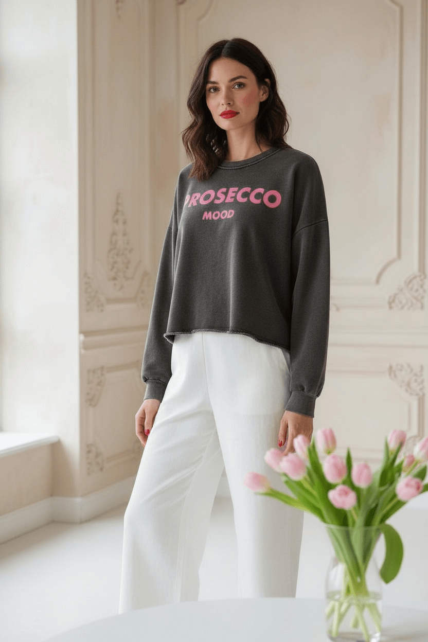 Frau trägt einen grauen Pulli mit der Schrift Prosecco Mode. Darauf dazu eine weiße Hose. Sie steht in einem hellen Raum mit Blumen auf dem Tisch.