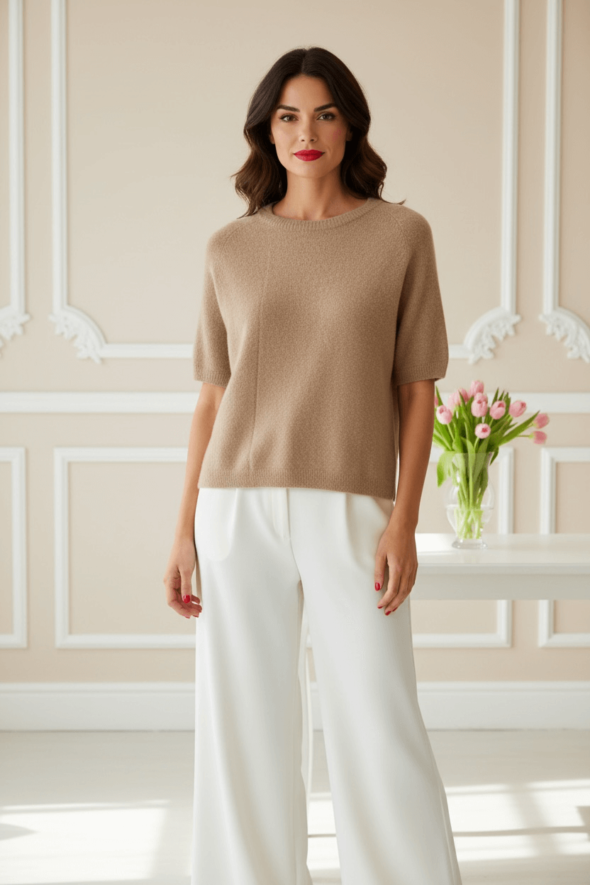 Frau steht in einem hellen Raum mit Blumen auf dem Tisch. Sie trägt einen beigen Pullover mit einer weißen Hose dazu.