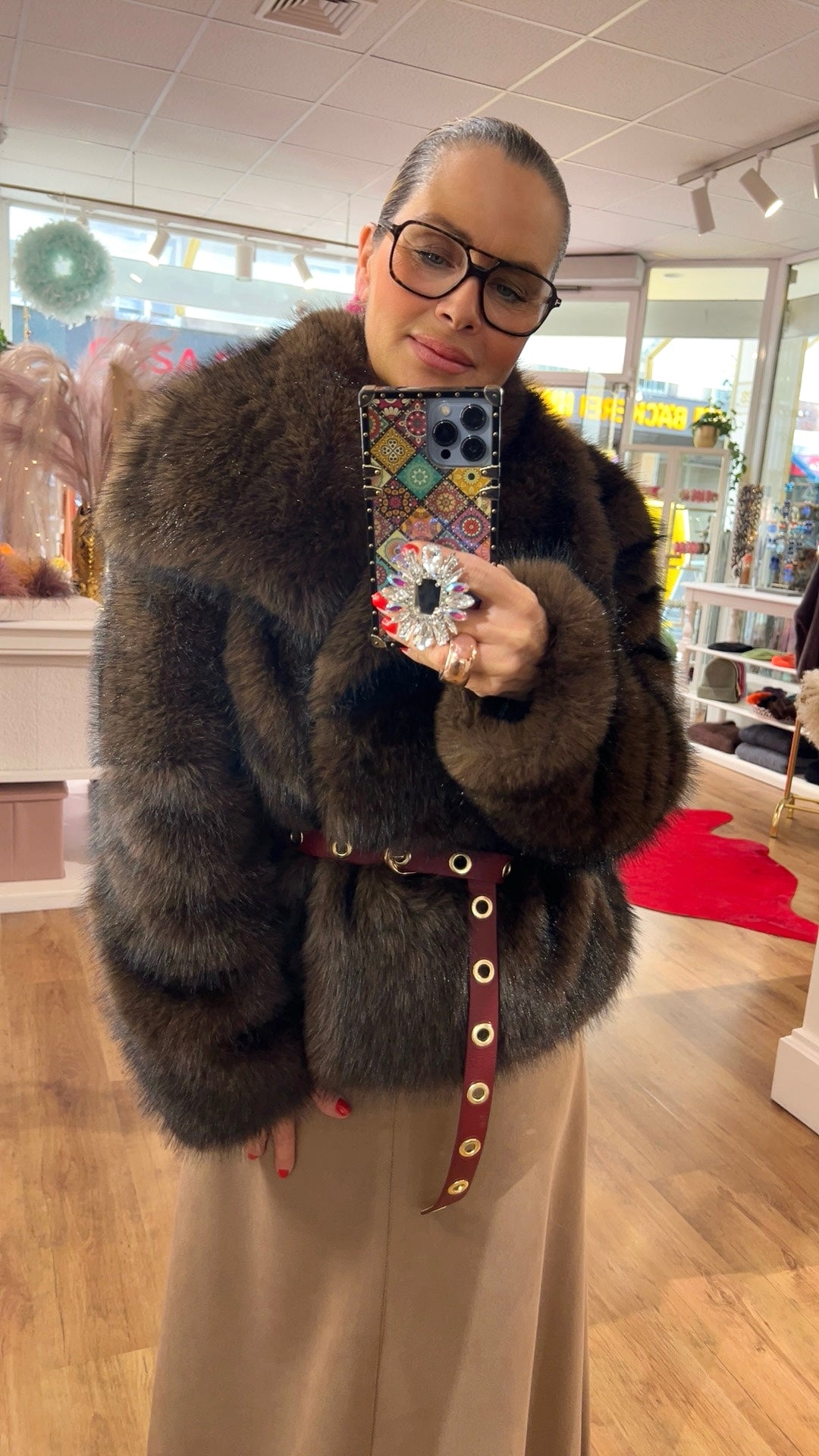 Person steht in einem Boutique-Innenraum und fotografiert sich im Spiegel. Sie trägt eine voluminöse, braun-pelzige Jacke mit breitem Kragen, einen burgunderfarbenen Gürtel mit goldenen Ösen und einen beigefarbenen Rock oder Hose. Die Person hält ein Smartphone mit bunter Hülle und großem, funkelndem Accessoire in der Hand. Im Hintergrund sind Regale, Modeartikel und Holzfußboden zu sehen.