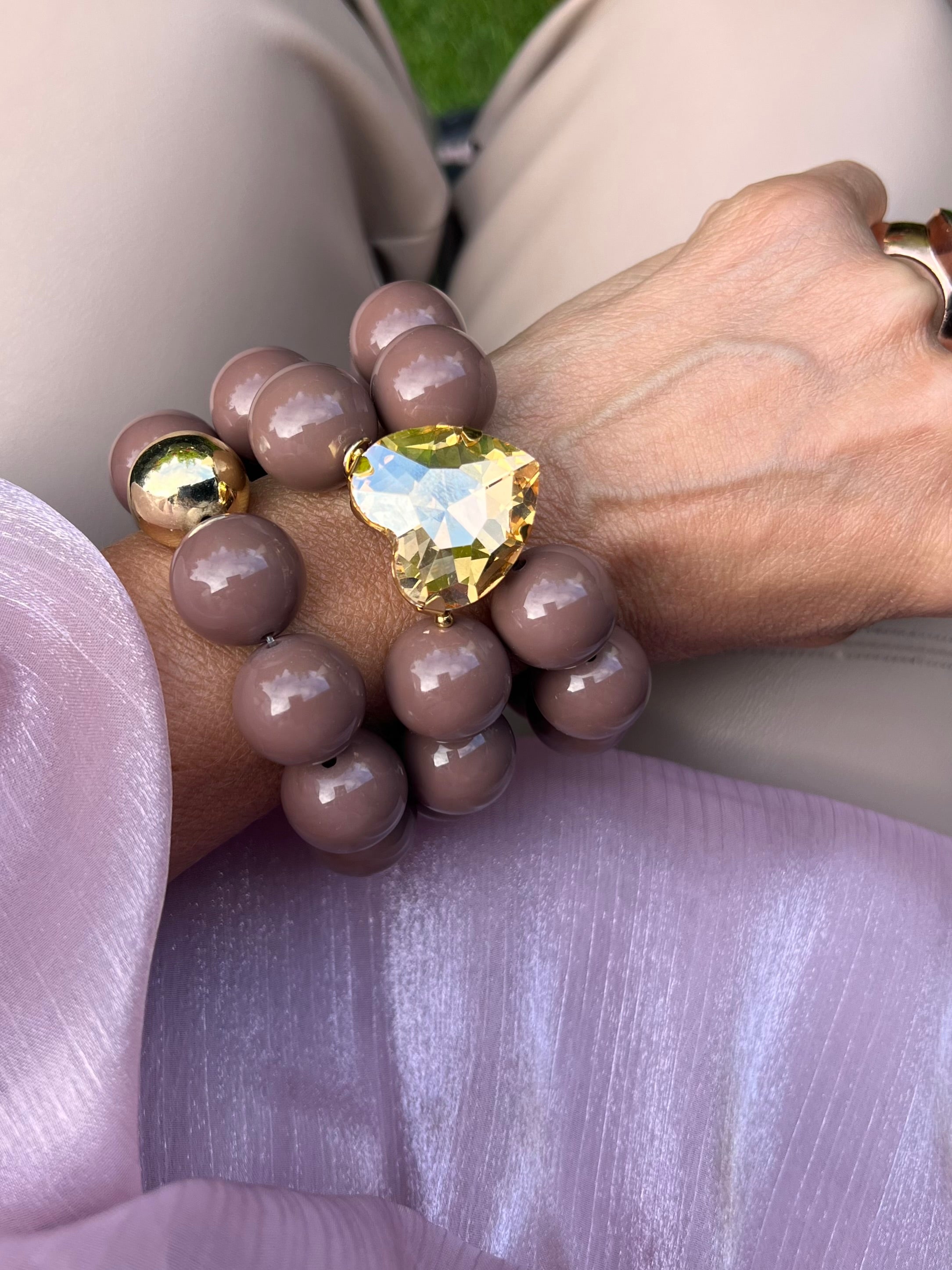 Zwei Perlenarmbänder in Taupe Farben mit großem Herzschmuckstein. Am Handgelenk auf rosa Stoff fotografiert.