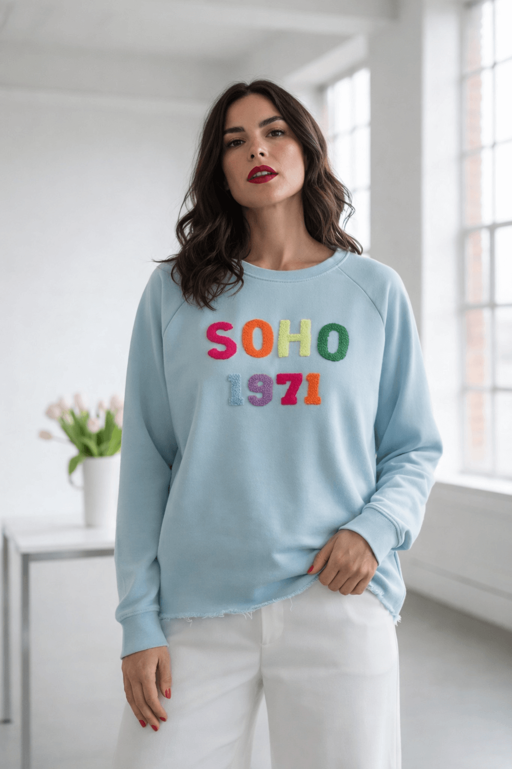 Frau trägt hellblauen SOHO 1971 Sweater mit buntem Schriftzug, lockerer Schnitt mit offenem Used-Look-Saum, kombiniert mit weißer Hose in hellem Raum mit Fenster und Tulpen.
