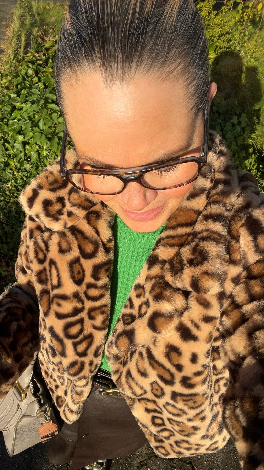 Nahaufnahme einer Person im Sonnenlicht. Zu sehen sind ein Mantel mit Leoparden Muster, ein grüner Strickpullover, eine Brille und eine beige Handtasche. Die Person blickt nach unten, der Hintergrund zeigt grüne Pflanzen.