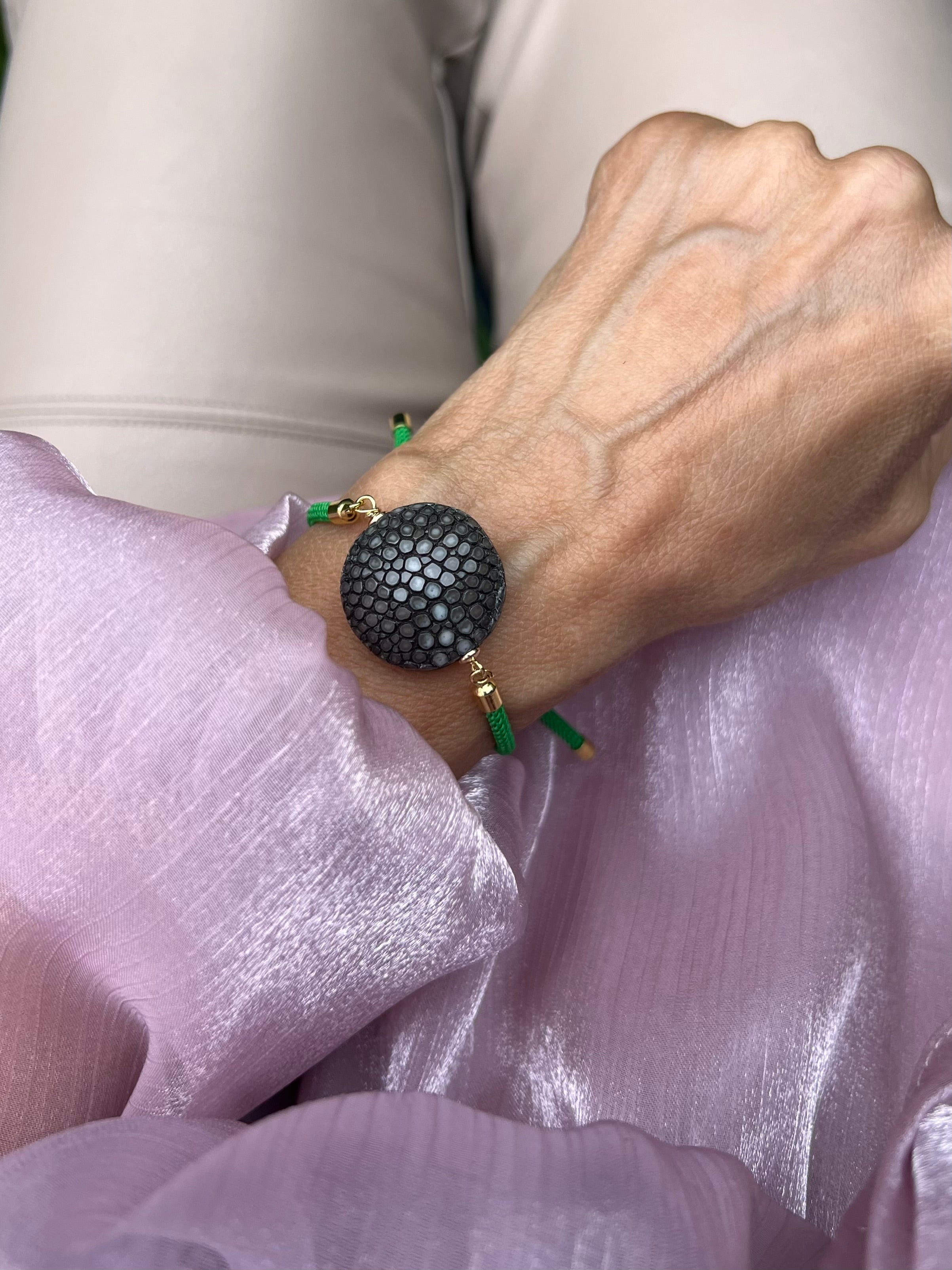 Grünes Armband mit brauner Perle und goldenen Details. Fotografiert am Handgelenk. Hintergrund rosa Stoff.