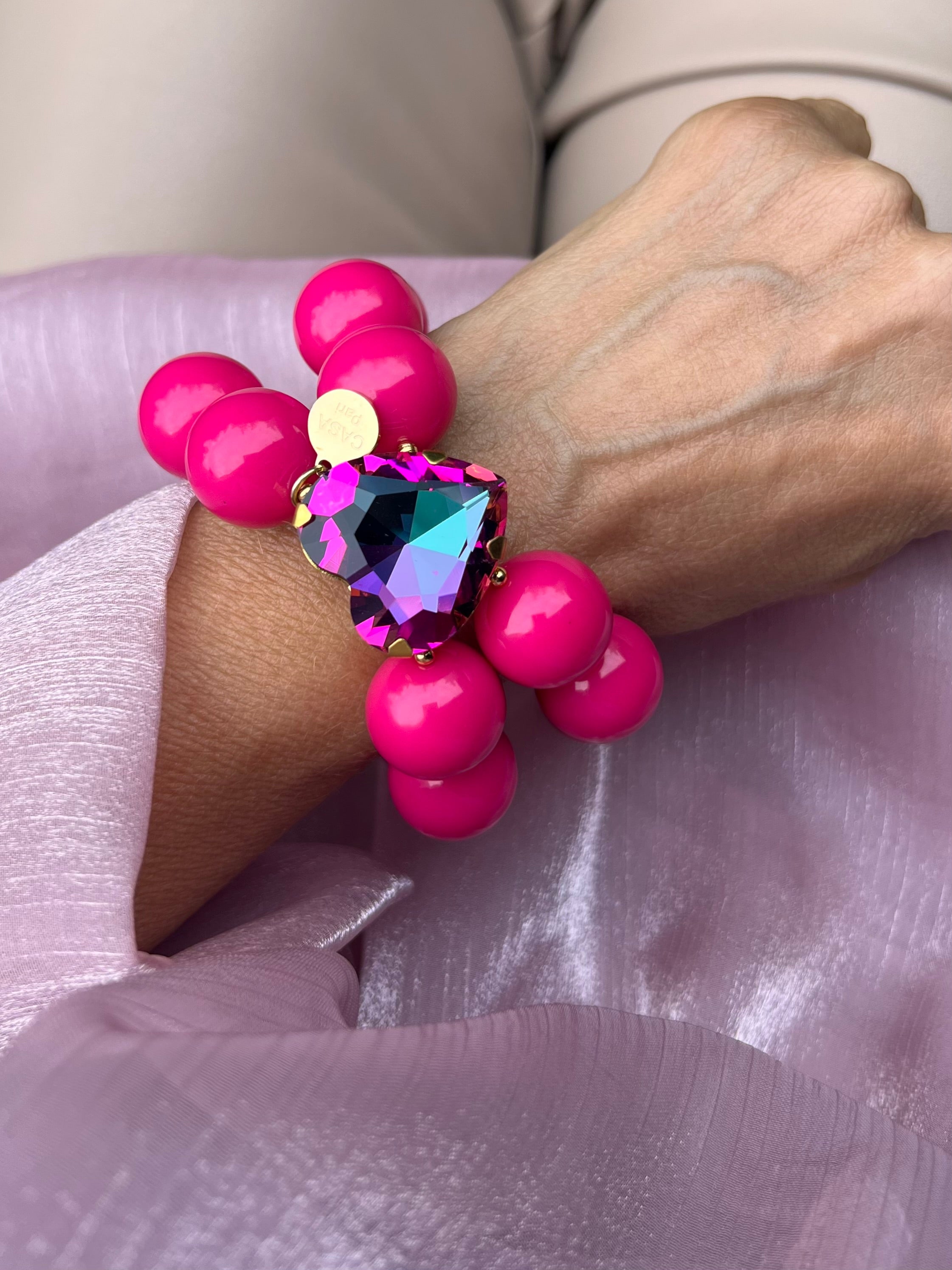 Handgelenk mit pinken Armband aus großen, glänzenden Perlen und einem herzförmigen, pink lila Kristall in der Mitte.