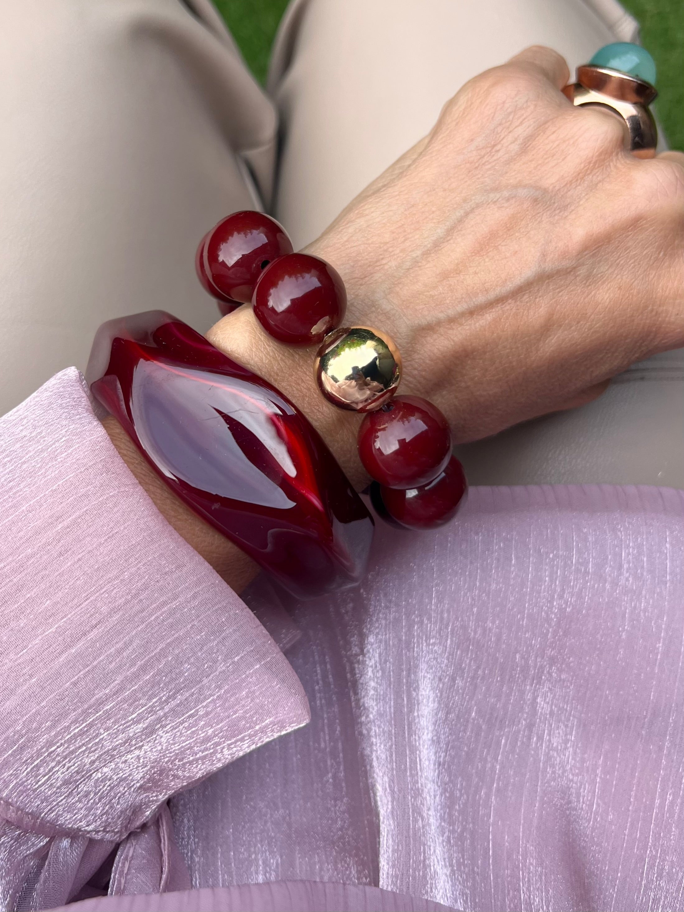 Zwei Armbänder in Burgunderfarben mit goldener Perle , großer Ring, am Handgelenk auf rosa Stoff fotografiert.