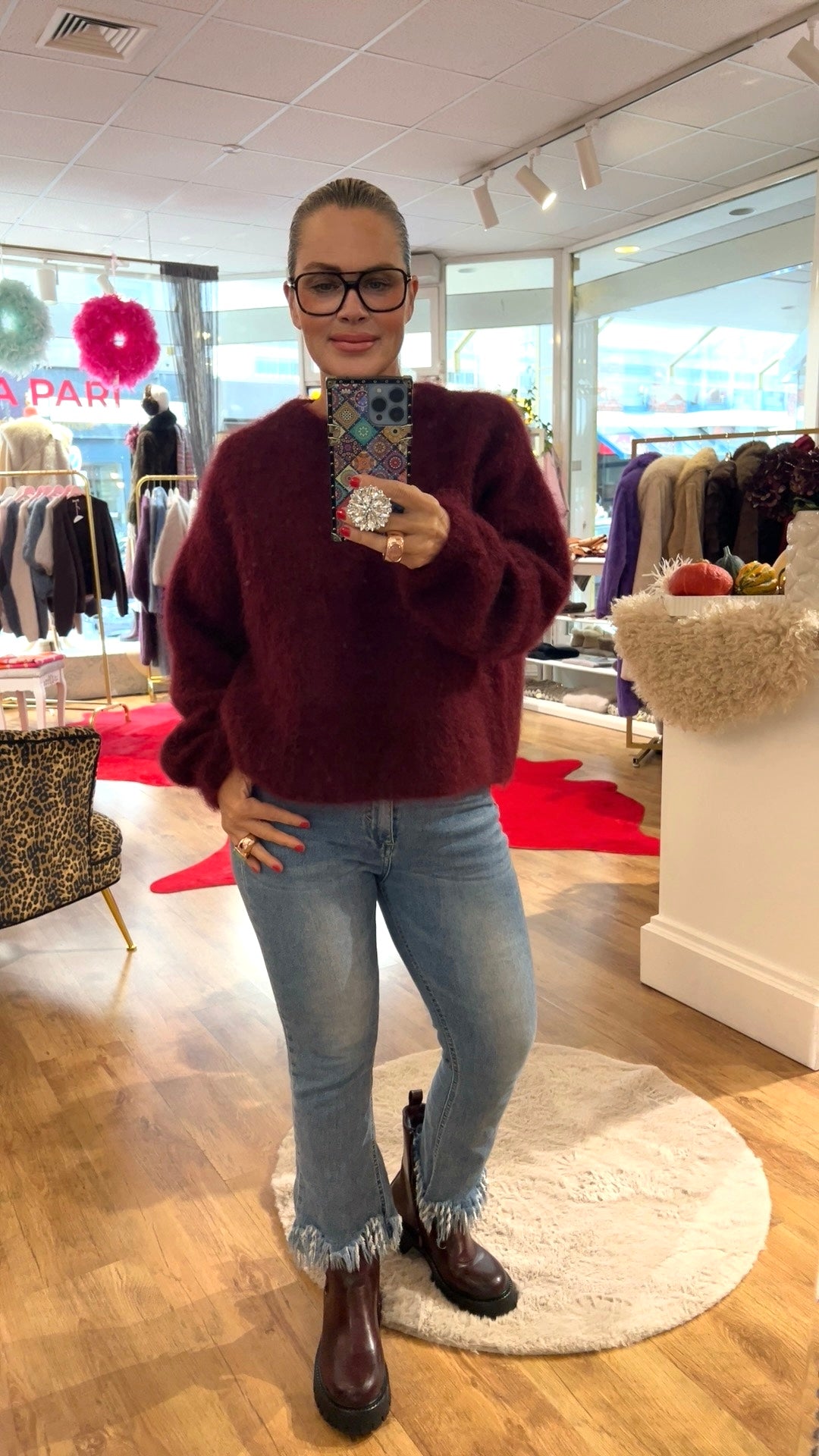 Frau trägt Bordeaux Farben, Wolle Pulli, dazu Jeans und braune Stiefel. Hält ein Handy in der Hand. Fotografiert in einer Boutique.