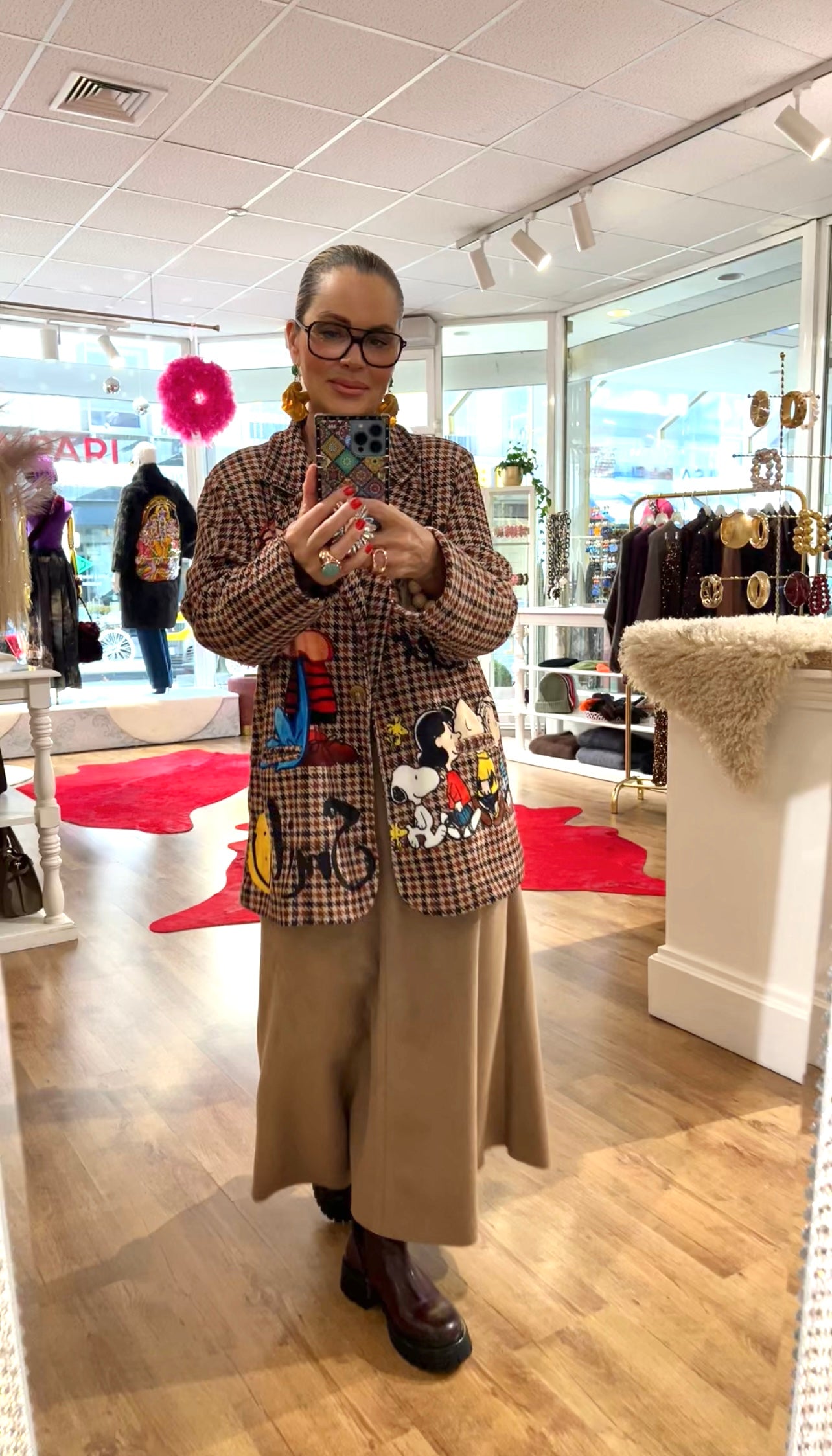 Person steht in einem Boutique-Store vor einem Spiegel und fotografiert sich mit dem Smartphone. Die Person trägt einen karierten Blazer mit bunten Comic-Motiven, ein bodenlanges beiges Rock- oder Hosen-Ensemble, dunkle Stiefel und auffällige Ohrringe. Im Hintergrund sind Kleidung, Accessoires und Dekorationen sichtbar.