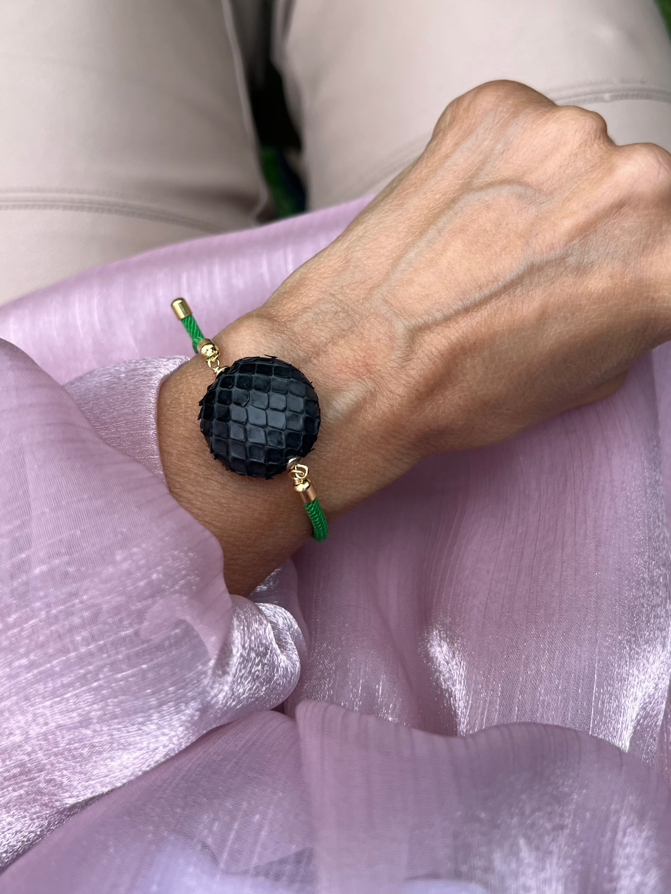 Grünes Armband mit schwarzer Perle und goldenen Details. Fotografiert am Handgelenk auf rosa Stoff.