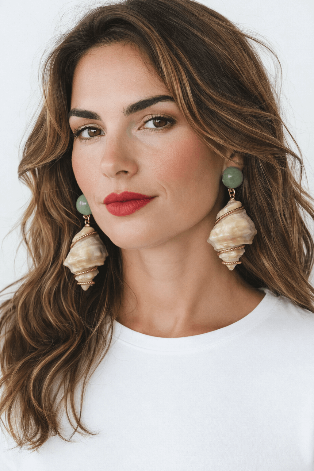 Frau mit braunen, welligen Haaren vor hellem Hintergrund, die grüne Clip-Ohrringe mit cremefarbenen Muschelanhängern und goldfarbenen Zwischenelementen trägt.