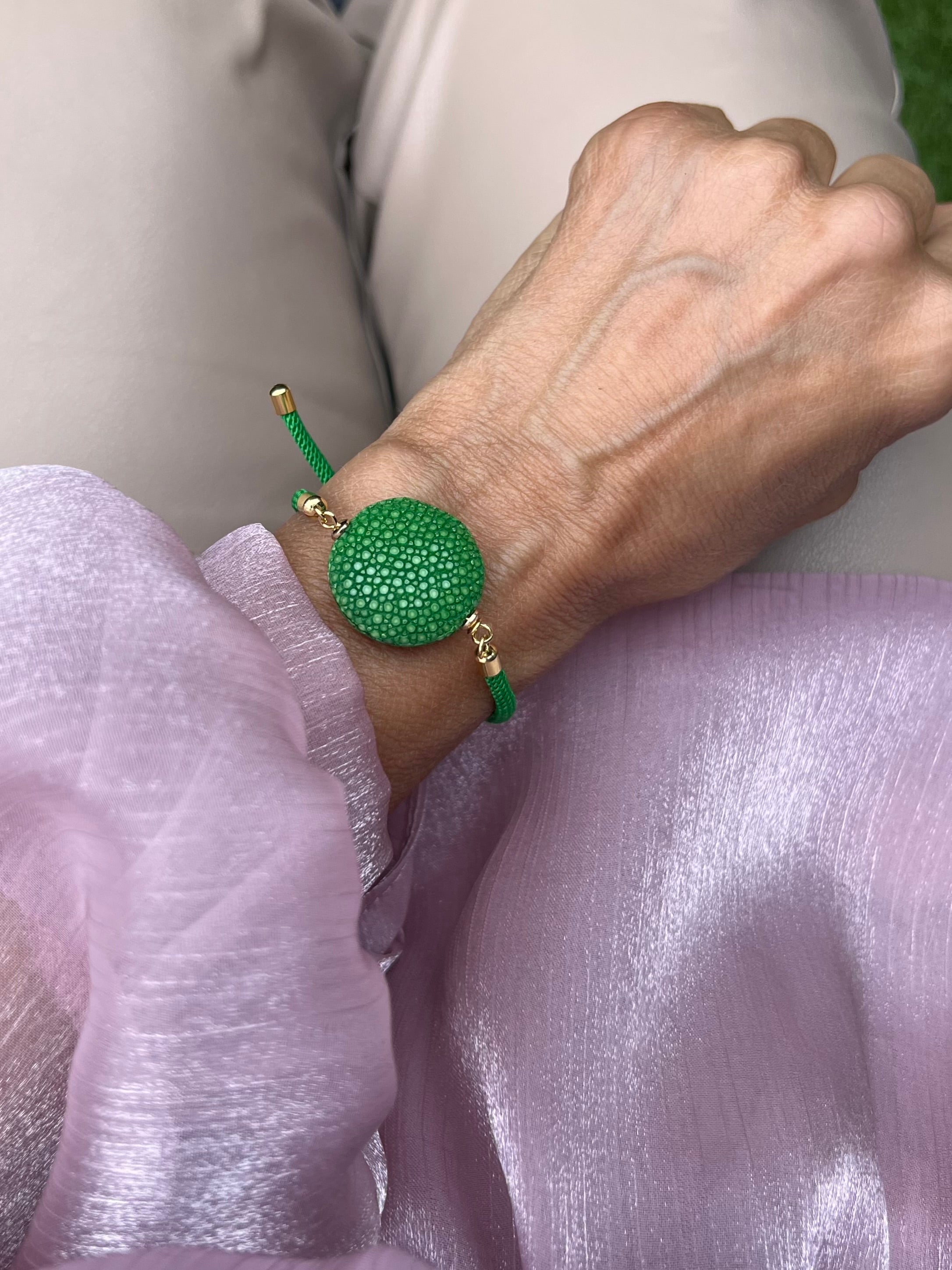 Grünes Armband mit grüner Perle und goldene Details. Am Handgelenk auf rosa Stoff fotografiert.