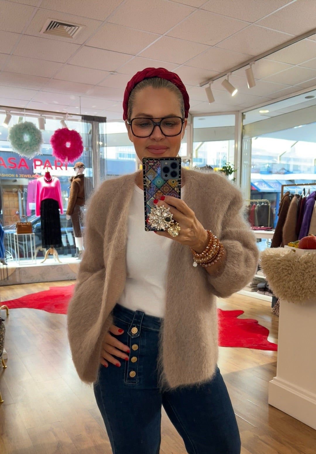 Frau trägt flauschige Strickjacke in beige, kombiniert mit weißem Shirt und dunkelblauer Jens mit Knopfleiste. Im Hintergrund bunte Mode und rote Akzente.