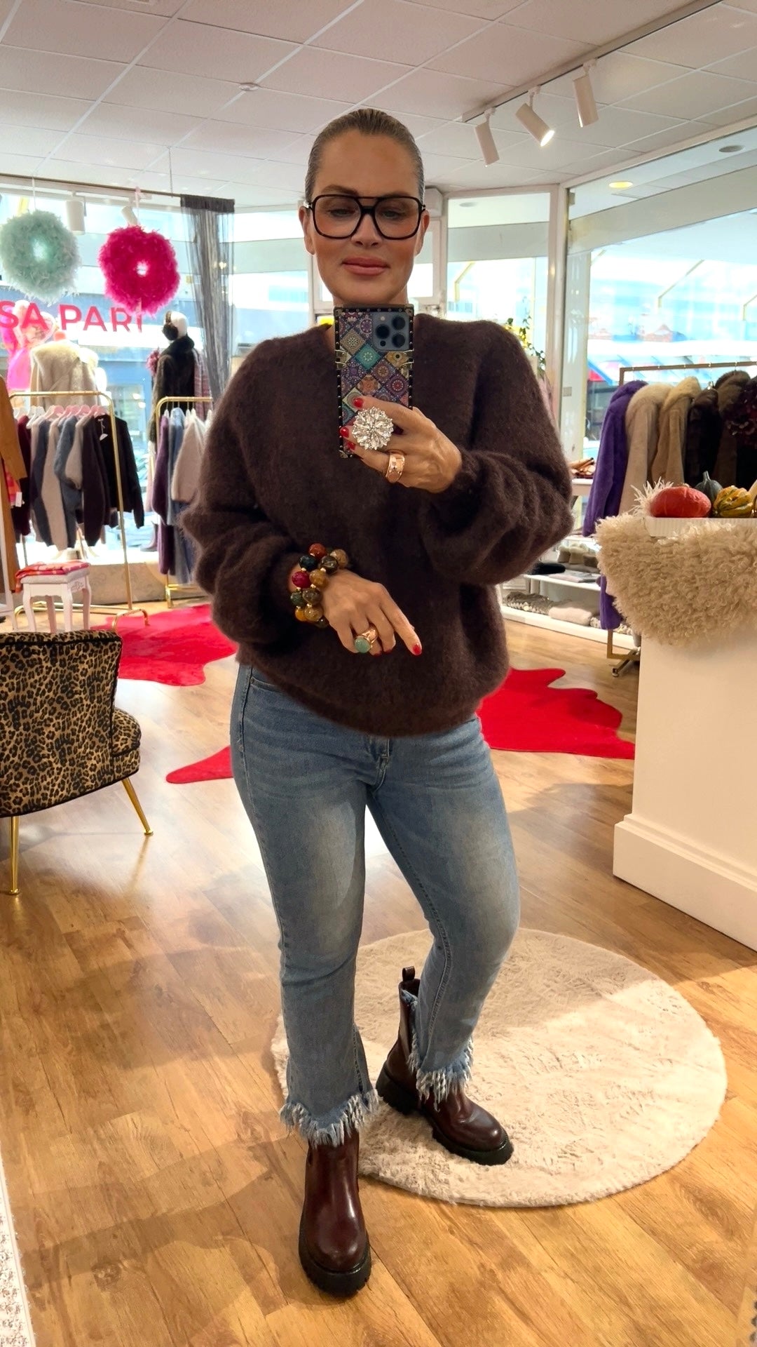 Frau trägt dunkelbraunen Mohairpulli, dazu Jeans und braune Boots. Hält ein Handy in der Hand fotografiert in einer Boutique.