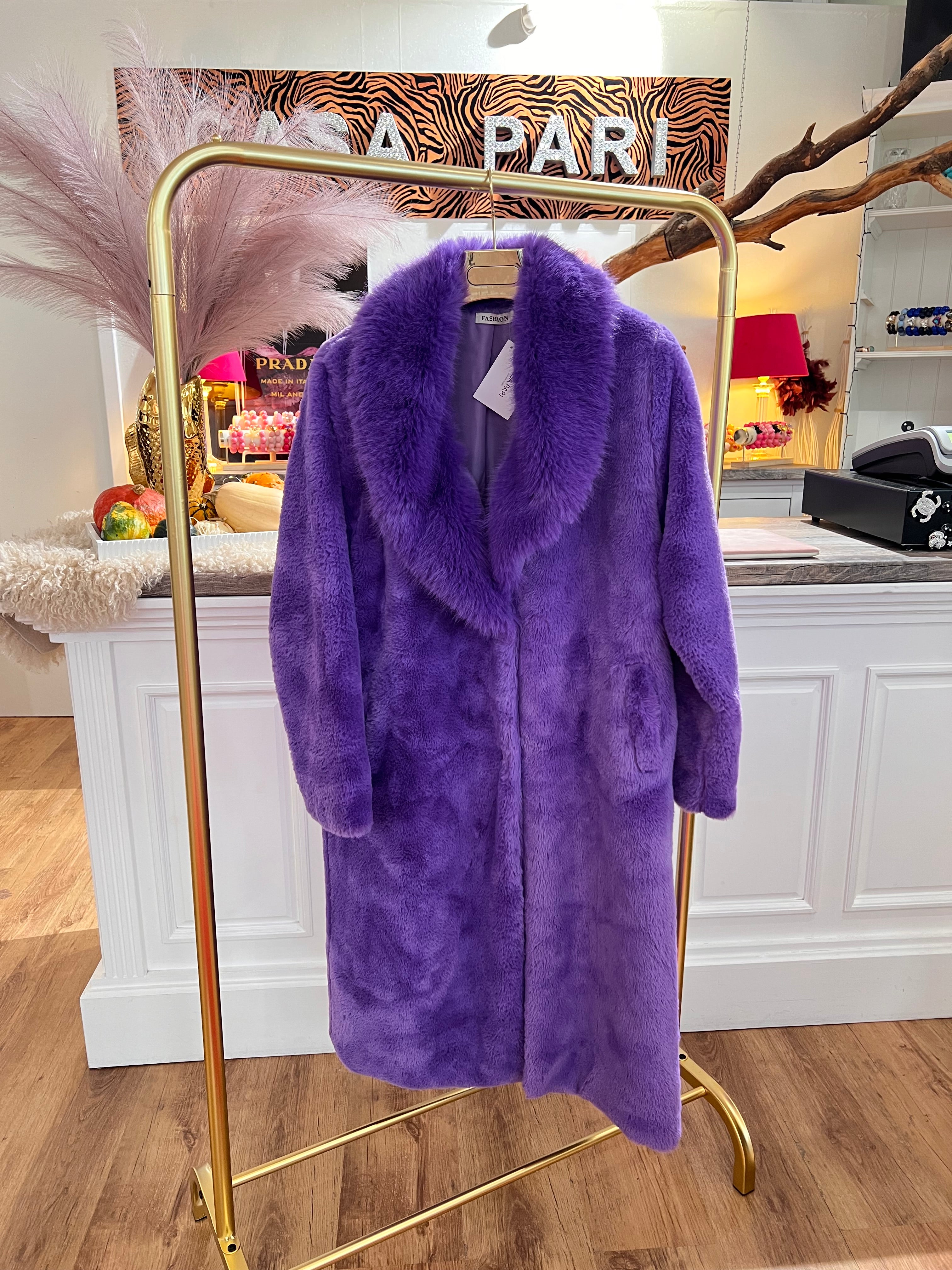 Langer, flauschiger Mantel in tiefen Violett hängt auf einem goldenen Kleiderständer vor einer weißen Theke.