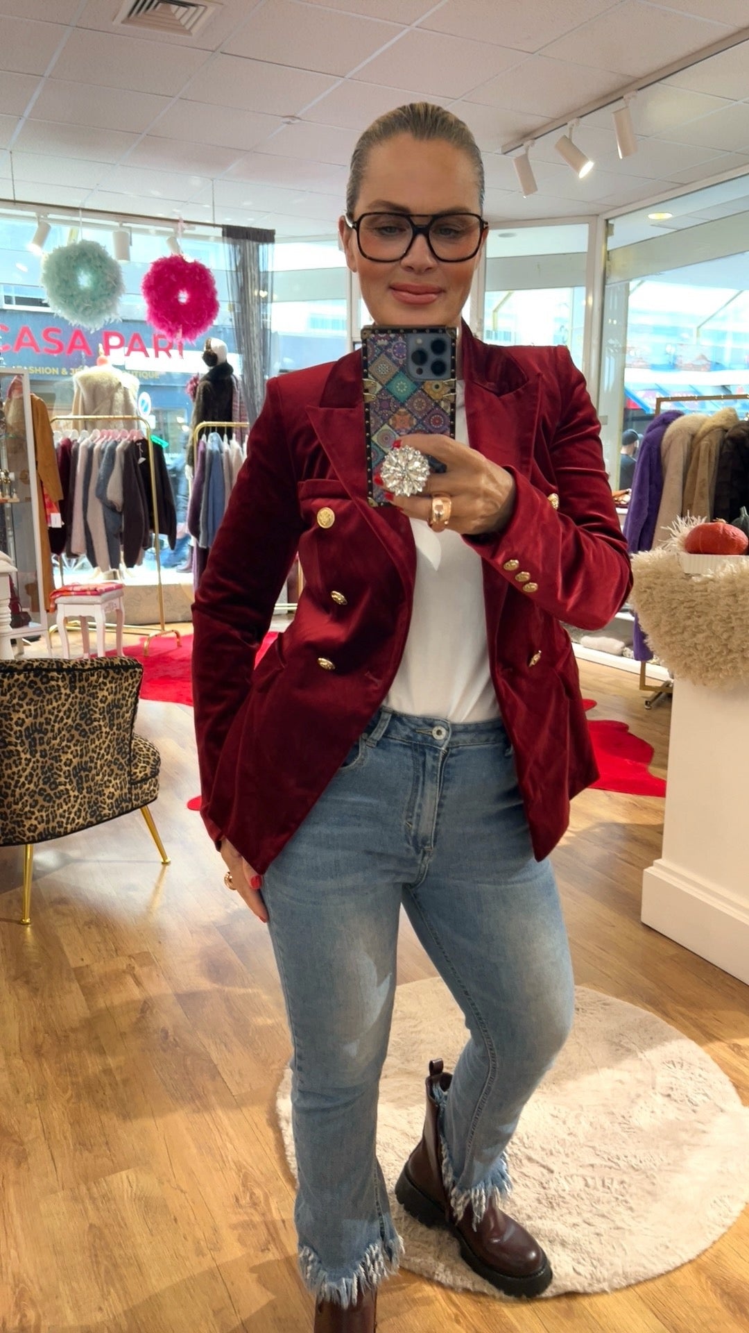 Frau trägt weinroten Samtblazer mit goldenen Knöpfen. Dazu eine Jeans und braune Boots. Sie hält ein Hand in der Hand. Fotografiere in einer Boutique.