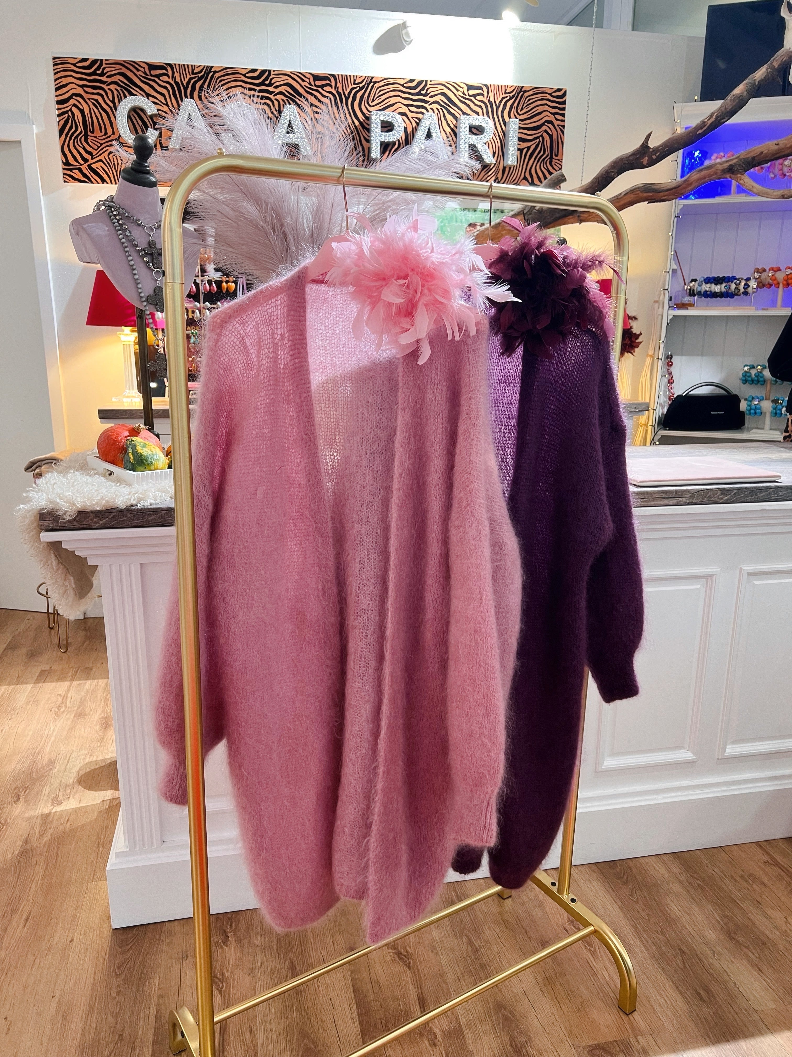 Zwei weiche Kid-Mohair Cardigans hängen auf rosafarbenen Bügeln an einer goldenen Kleiderstange in einer Boutique. Ein in hellem Rossa, der andere in dunklem Violett. Beide haben lange Ärmel und eine offenen Front. Blumen am Kragen.