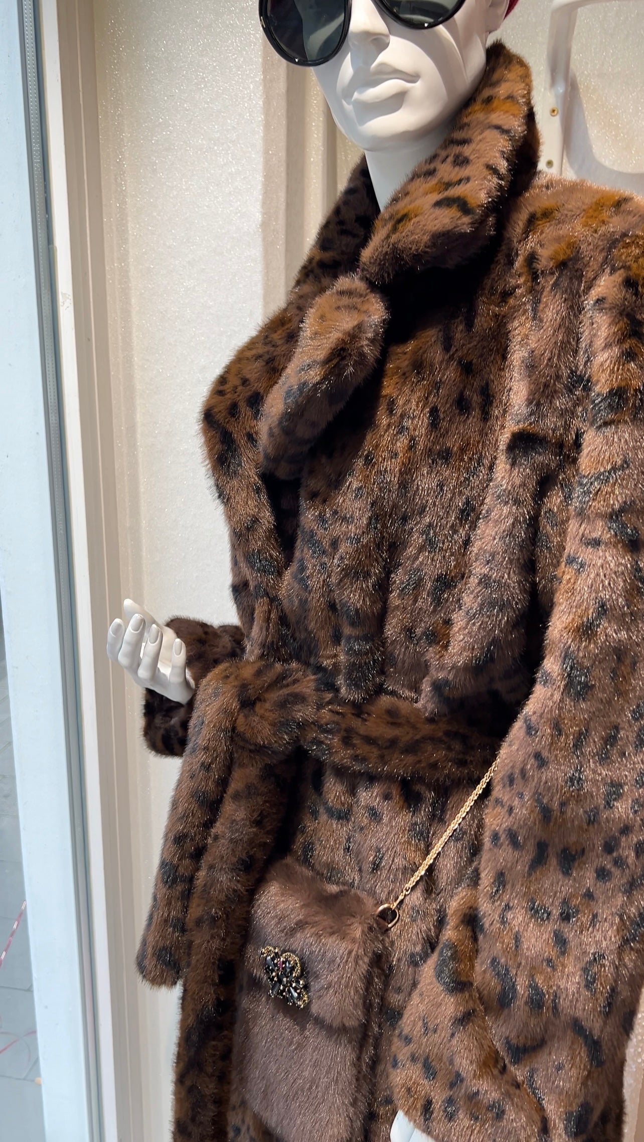 Brauner Kunstpelzmantel mit Leopardenmuster auf einer Schaufensterpuppe, dazu eine kleine Umhängetasche mit goldener Ketten und Brosche.