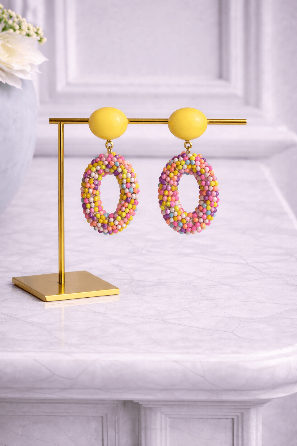 Ovale Ohrringe mit gelbem Clip-Verschluss und bunten Perlen in Pastelltönen, präsentiert auf goldenem Schmuckständer vor hellem Hintergrund.