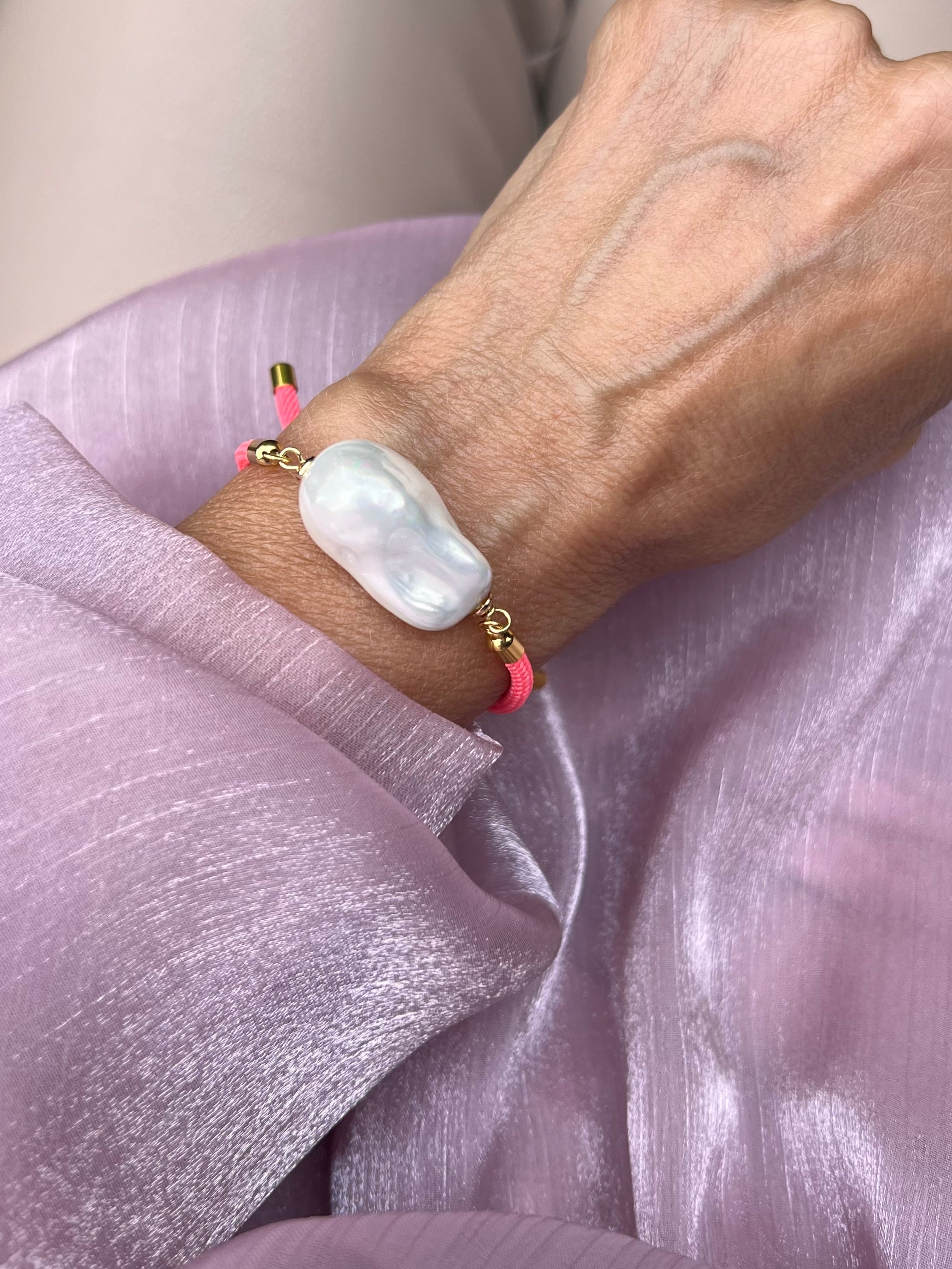 Handgelenk mit feinem korallefarbenem Armband und einer Barockperle mit goldenen Details. Auf rosa Stoff fotografiert.