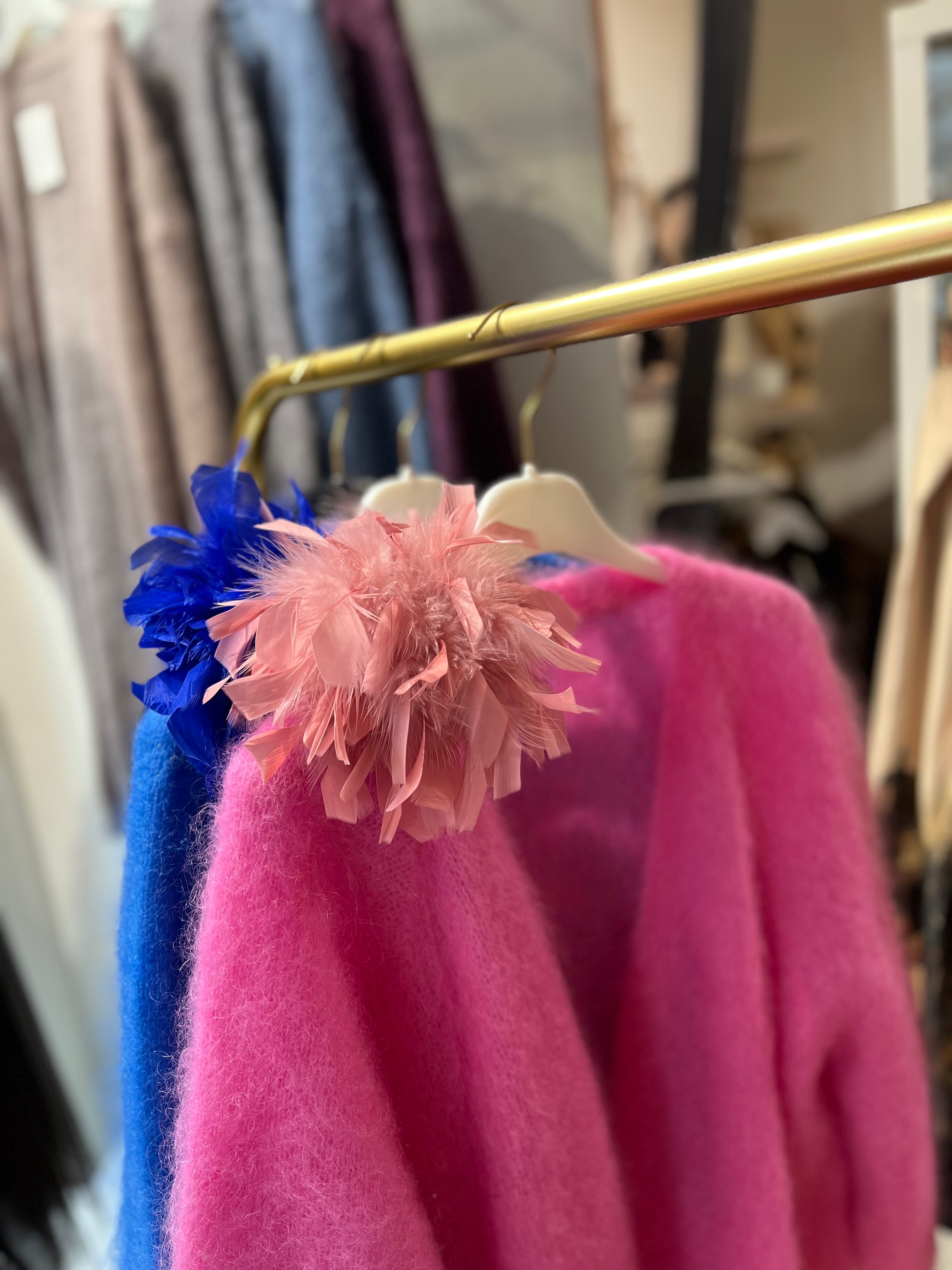 Zwei Federbroschen mit großen Blüten aus federn, einer in Rosa und einer in leuchtendem Blau. befestigt an einem pinkfarbenen Pullover auf einem goldenen Kleiderständer.