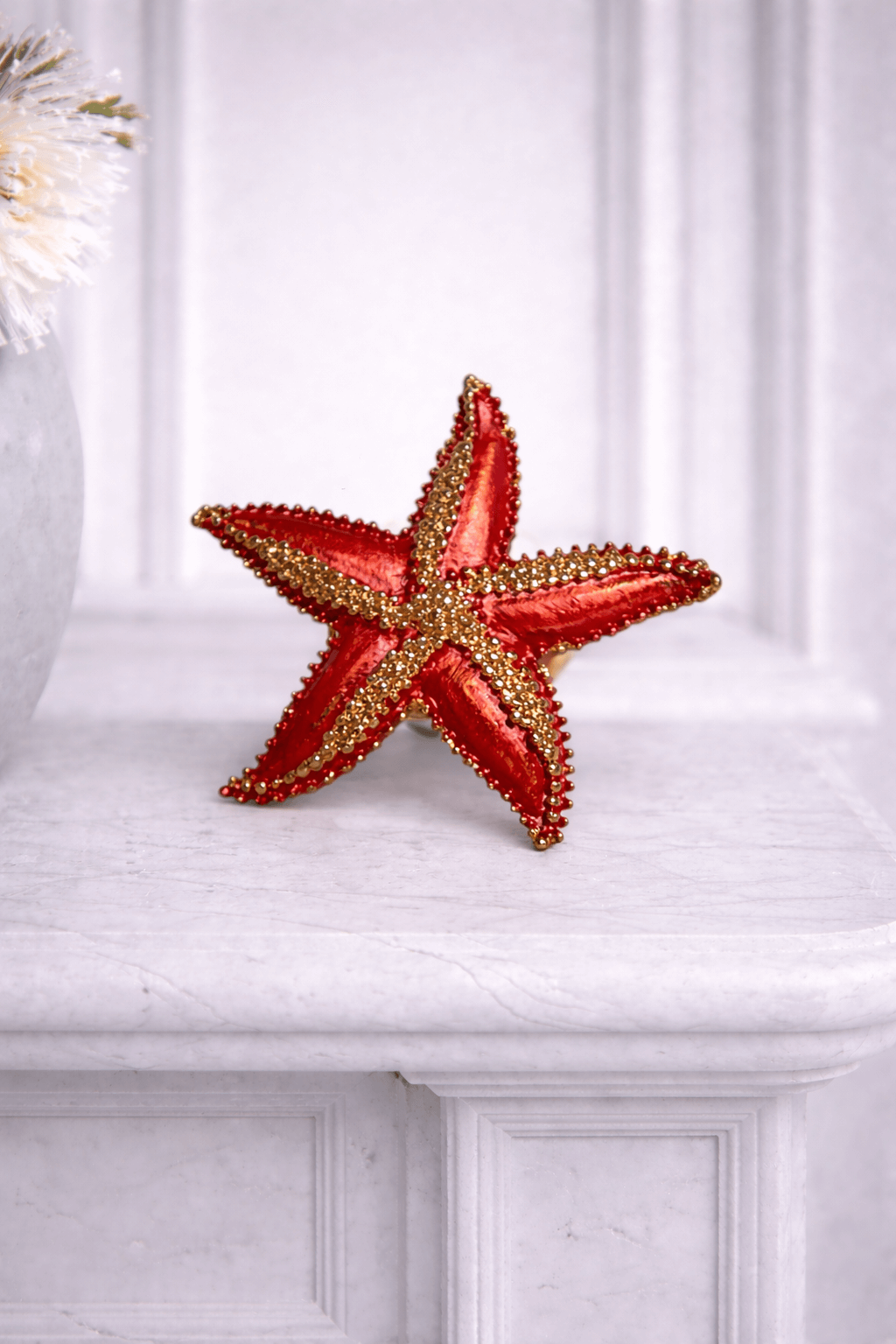 Rote Seestern-Brosche mit goldfarbenen Details auf heller Marmorplatte vor weißem Hintergrund.
